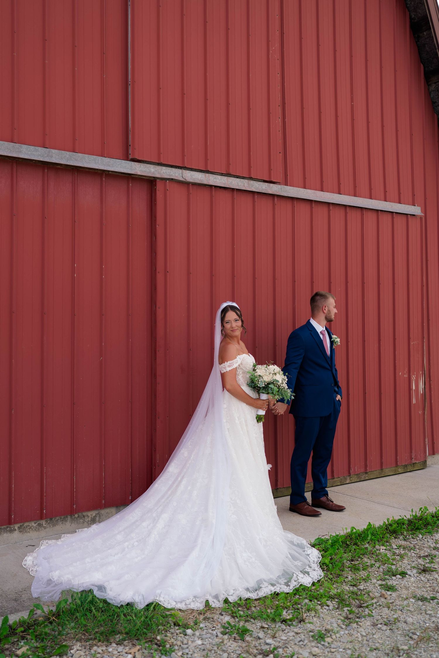Bride in white lace gown with long veil stands with groom in navy suit before a red barn wall.