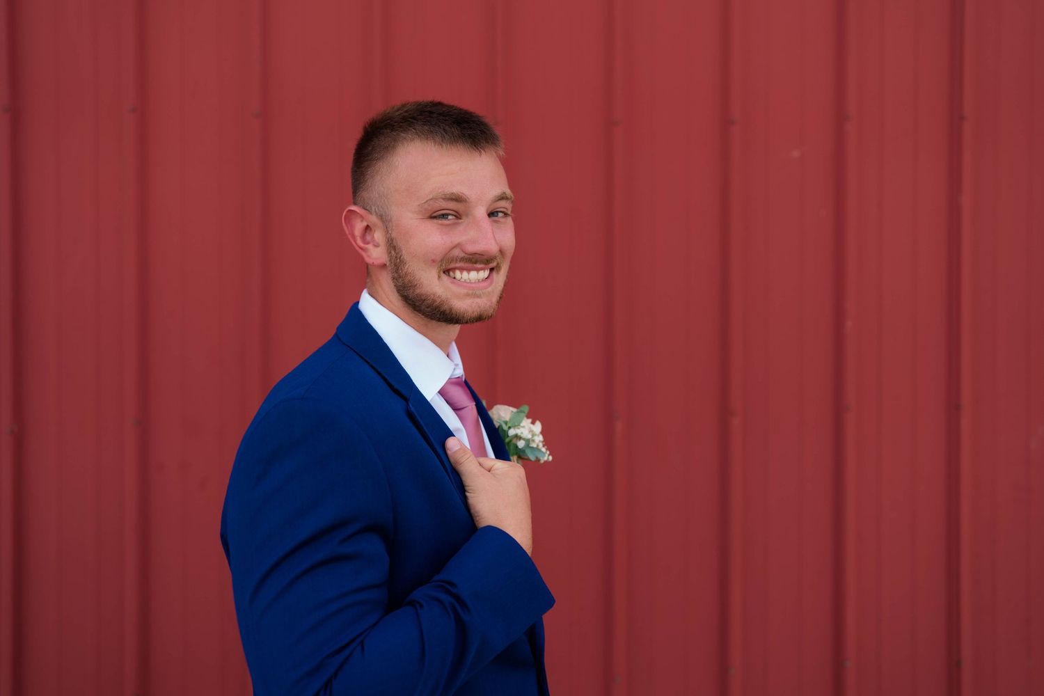 Smiling groom in navy suit with pink tie and floral boutonniere posing against red barn wall.