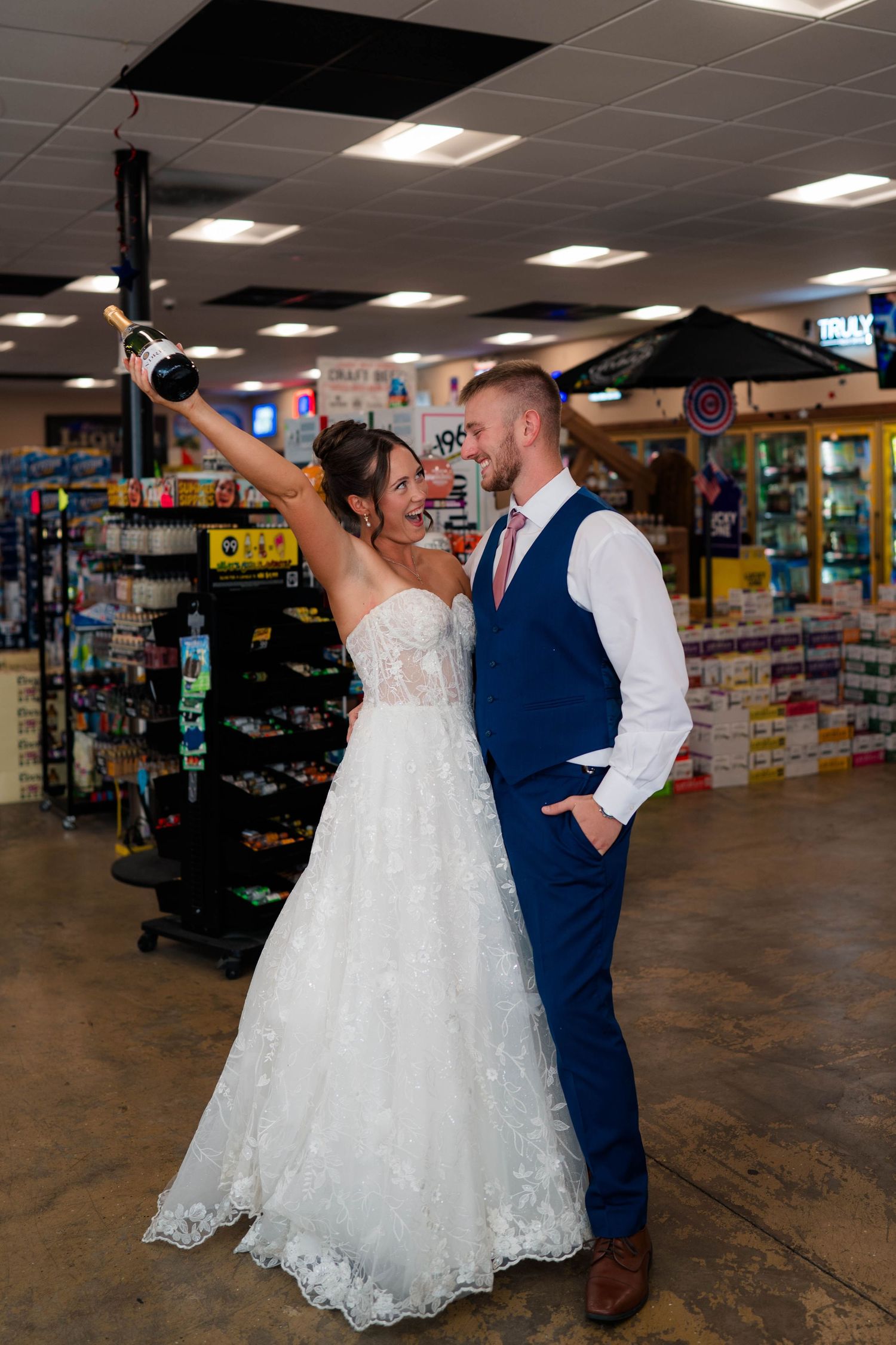 Happy bride and groom celebrating in convenience store, bride raising champagne bottle in wedding attire.