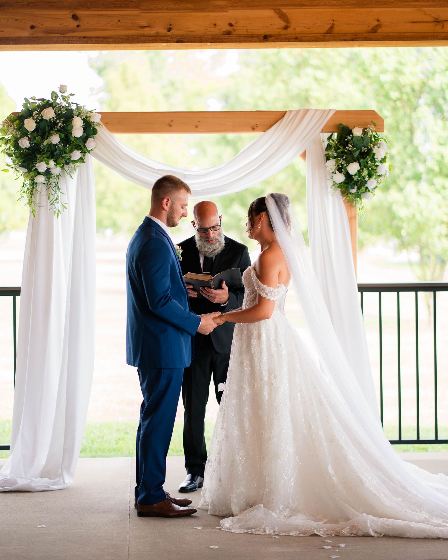 Bride and groom exchange vows under wooden arch with white draping and floral arrangements at ceremony.
