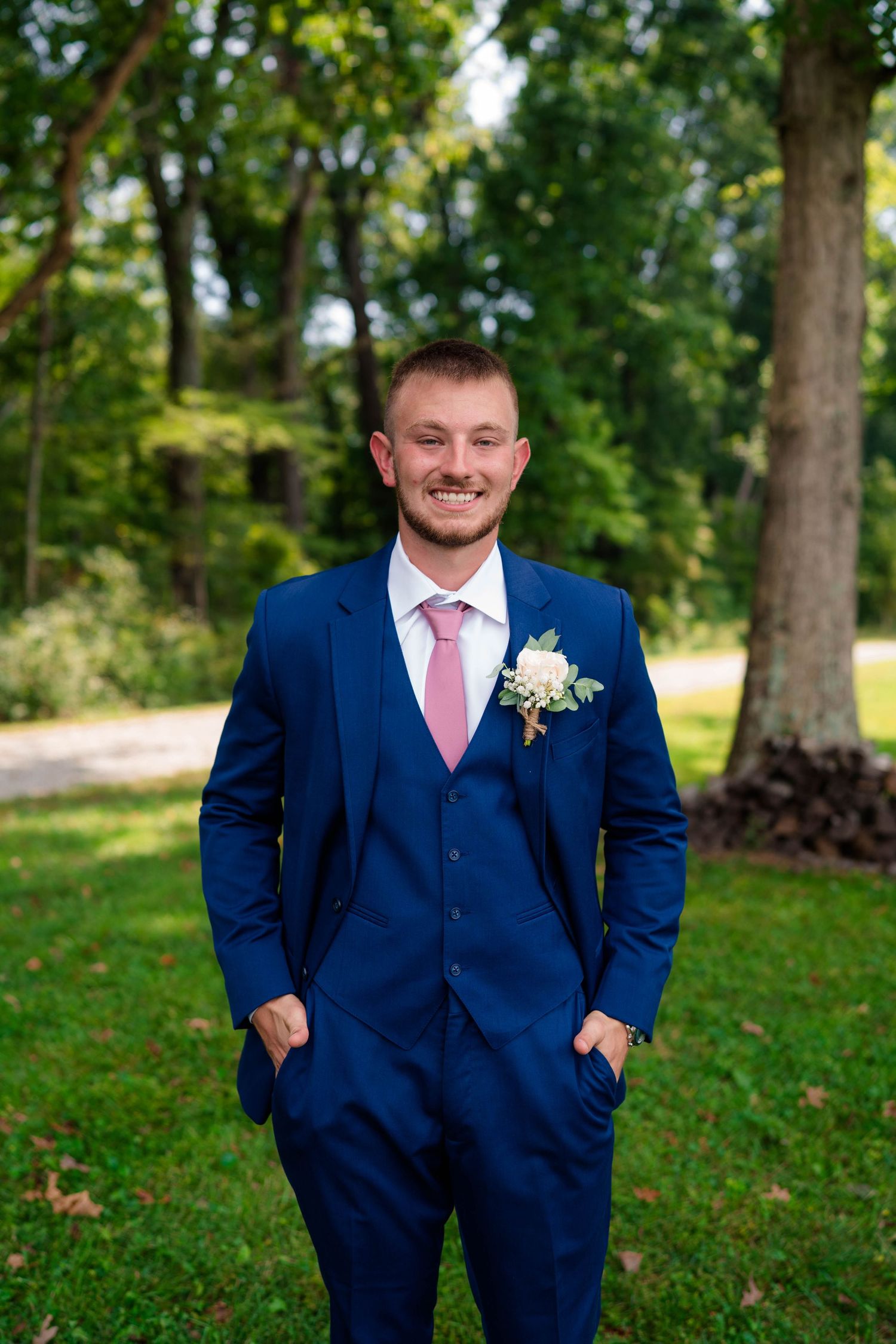Smiling groom in navy blue three-piece suit with pink tie and floral boutonniere standing outdoors.