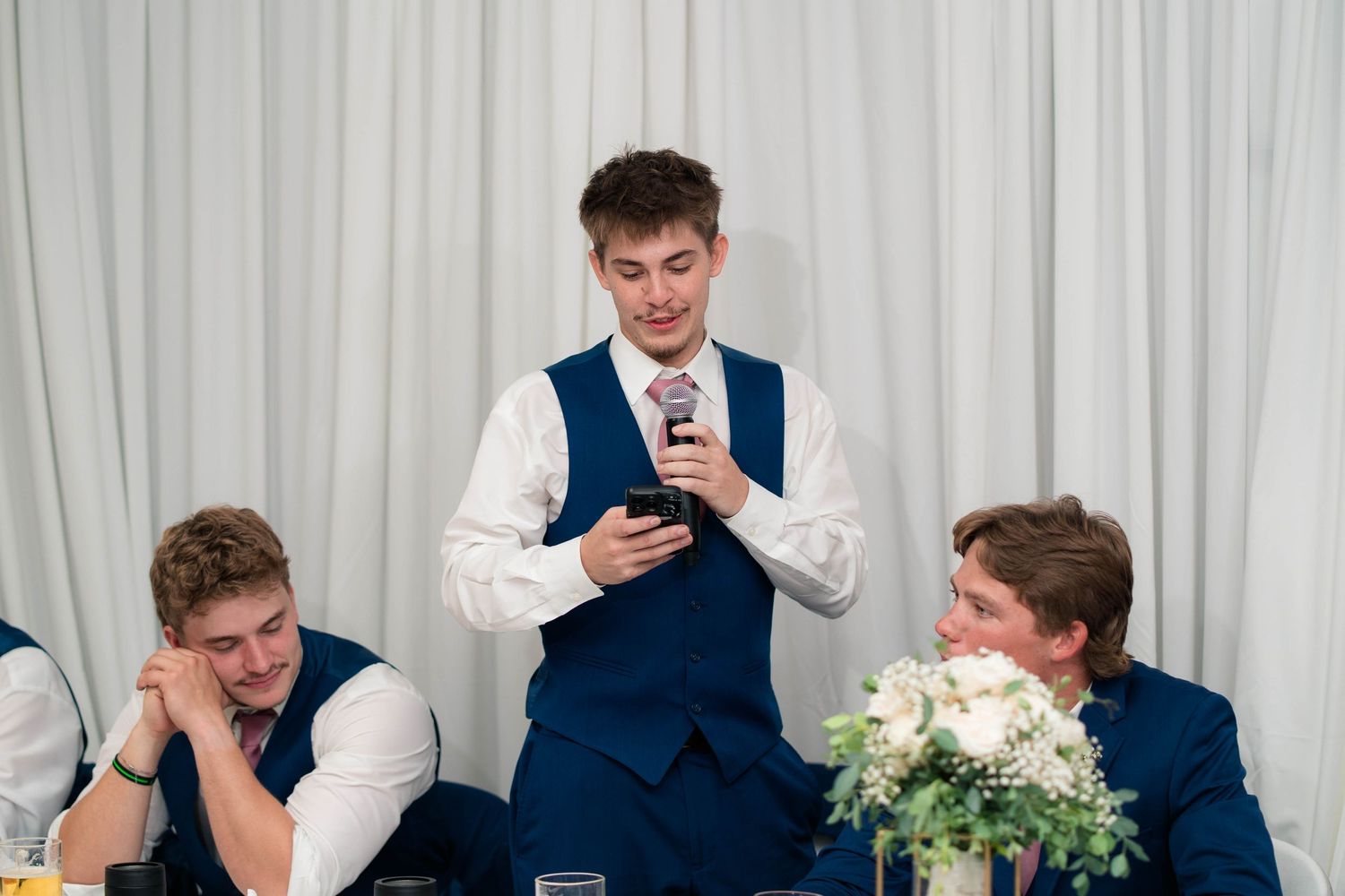 Groomsman reads wedding speech from phone while seated groomsmen listen at decorated reception table.