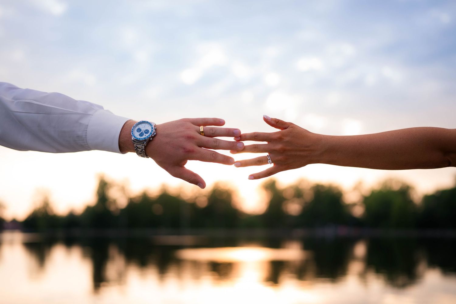 Bride and groom reach for each other's hands near a reflective lake at sunset, showing wedding rings.