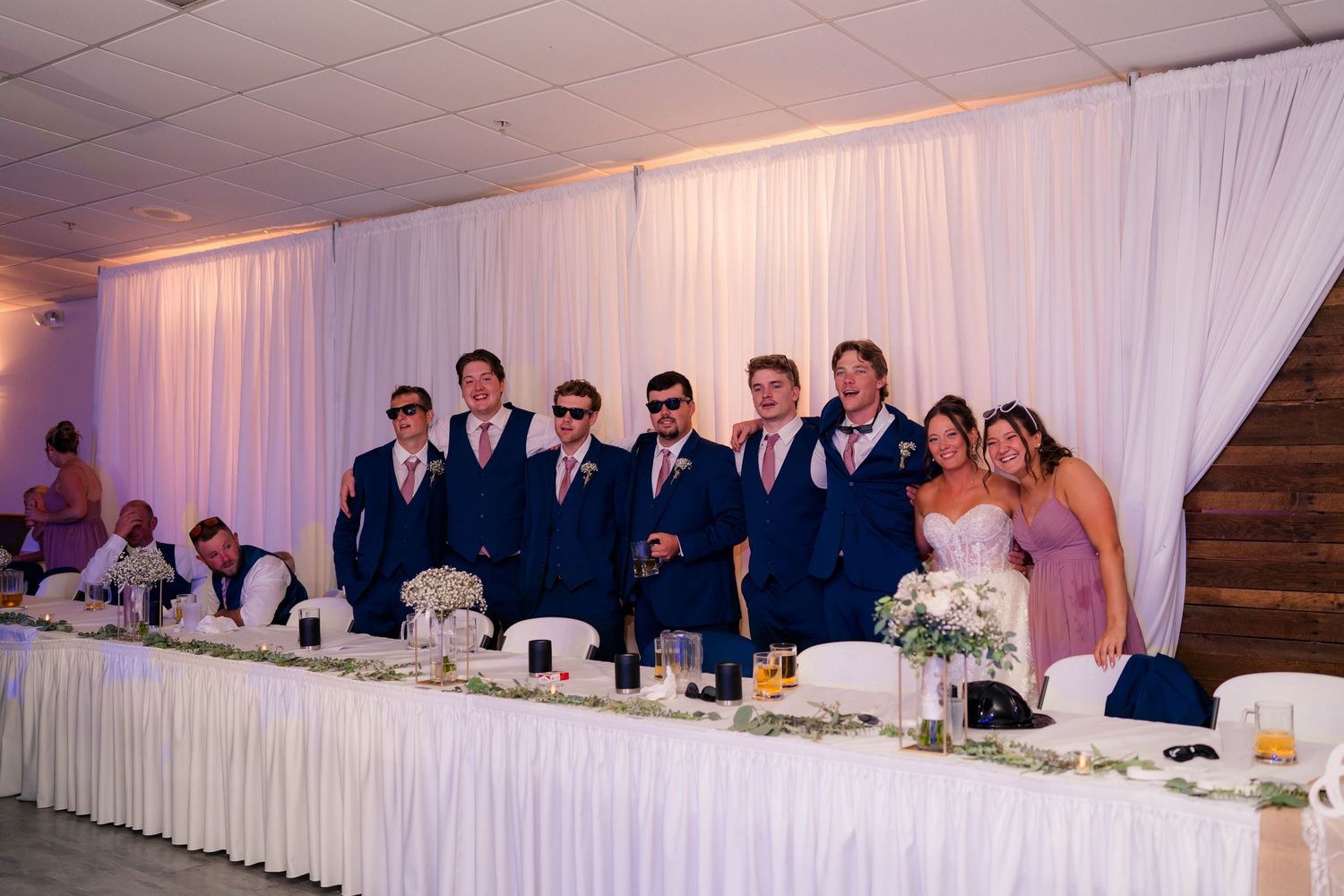 Groomsmen in navy suits and pink ties pose with sunglasses alongside the wedding party at the reception.