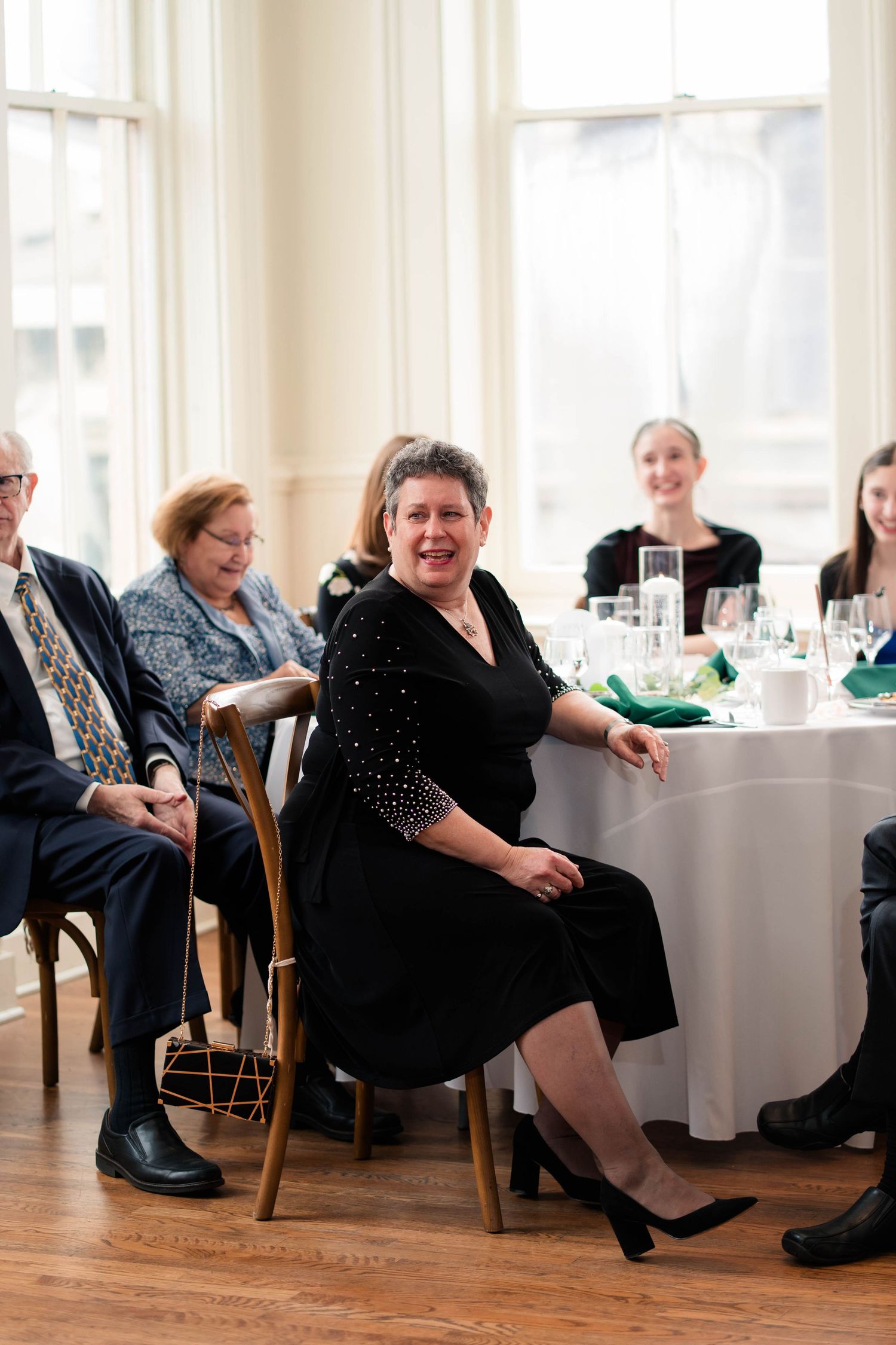 Woman in black dress smiling at wedding reception seated at table with other guests nearby.