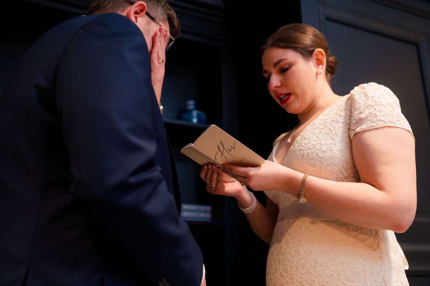 Bride in lace gown reading handwritten wedding vows from small booklet during private ceremony.