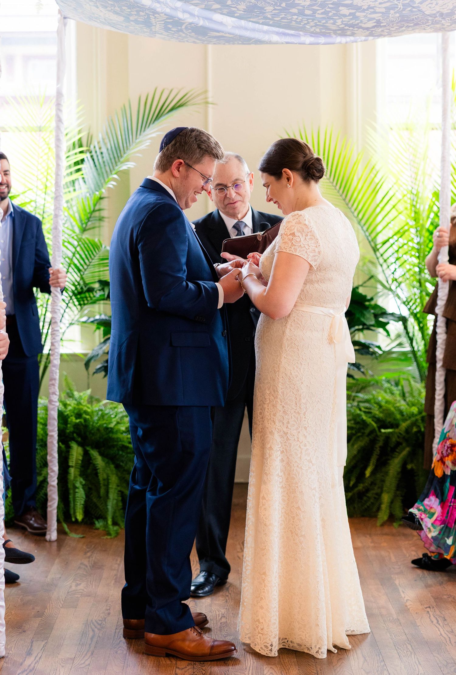 Groom places wedding ring on bride's finger under chuppah with palm leaf decorations during Jewish ceremony.
