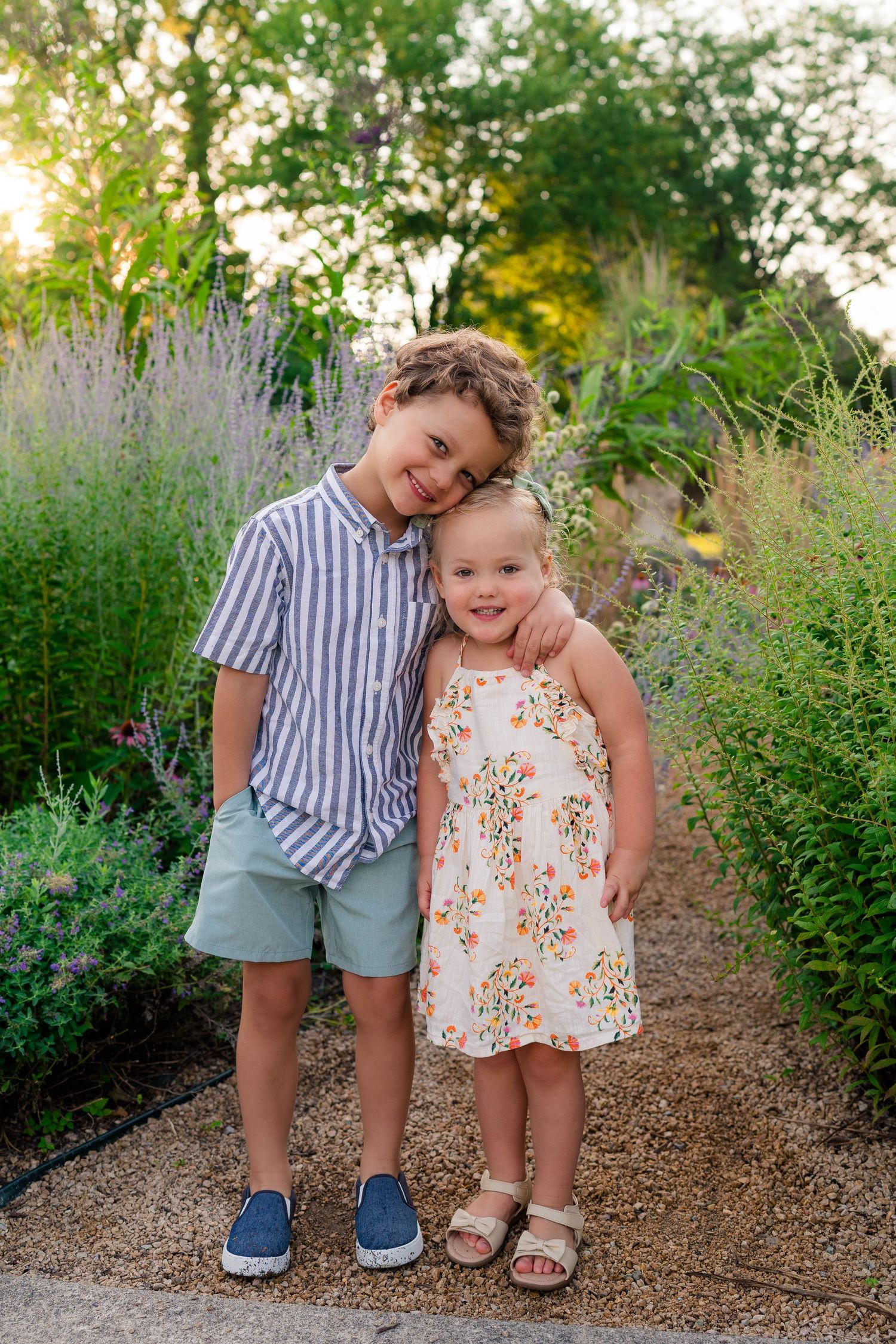 Two young siblings pose together on a garden path surrounded by lush greenery and wildflowers at golden hour.