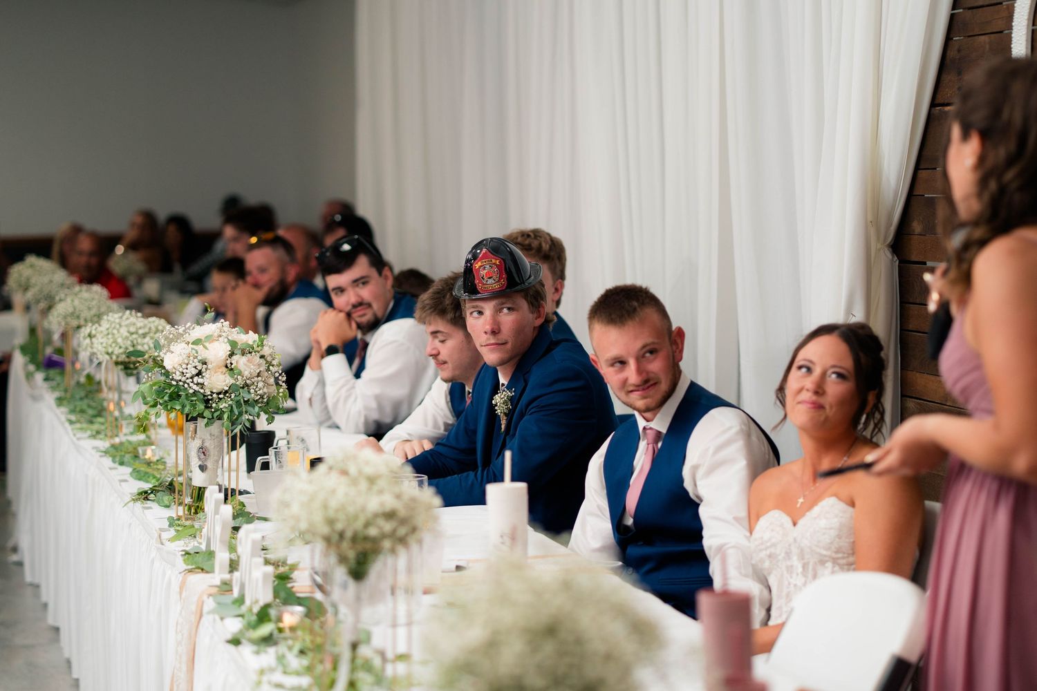 Wedding party and guests seated at a long decorated head table with white floral centerpieces at reception.