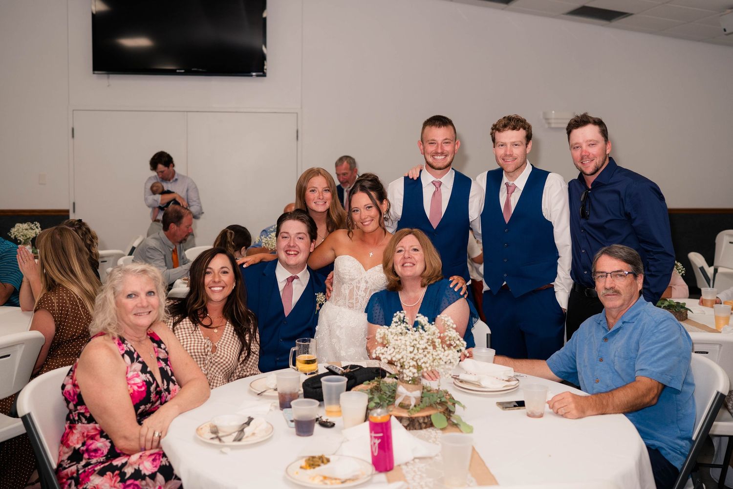 Wedding reception group photo with bride and groom surrounded by smiling family and friends at a decorated round table.