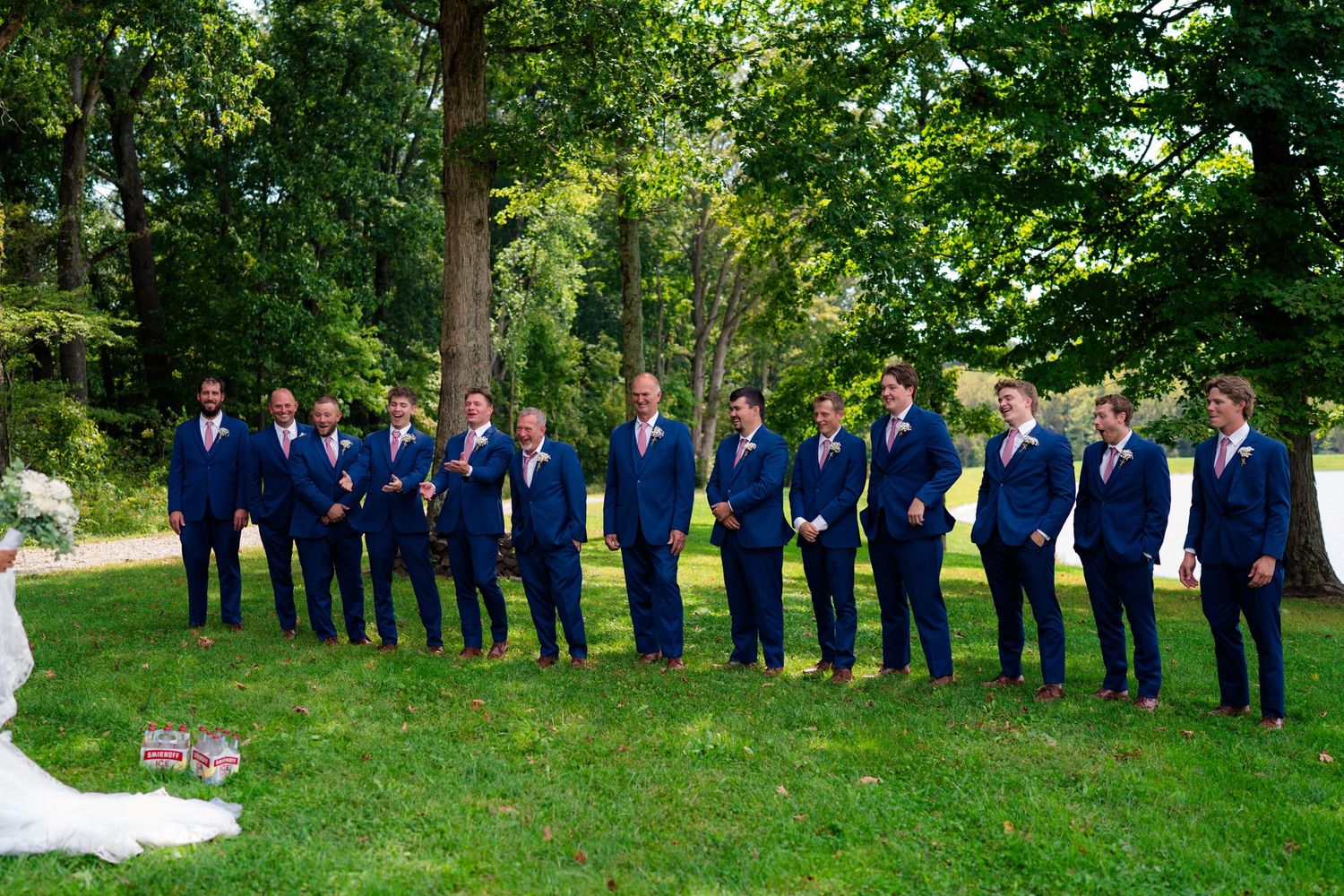 Large wedding party of groomsmen in matching navy suits with pink ties lined up outdoors on green lawn.