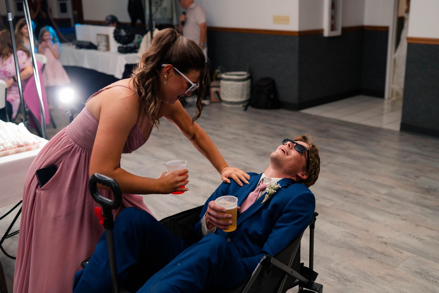 Bridesmaid in pink dress wearing novelty glasses leaning over a groomsman in a navy suit at a wedding reception.