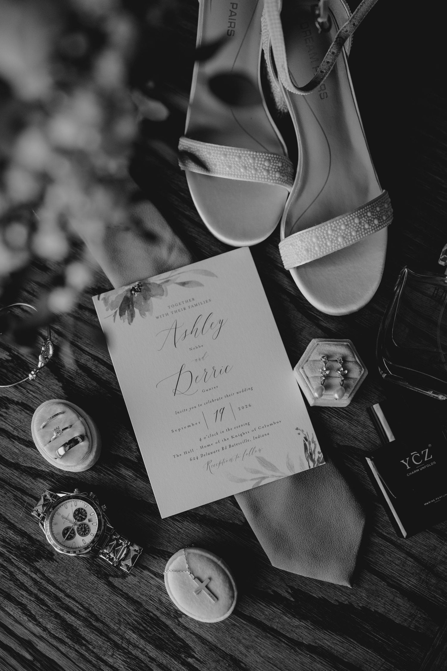 Black and white flat lay of wedding invitation for Ashley and Derrie surrounded by shoes, rings, watch, tie, and accessories.