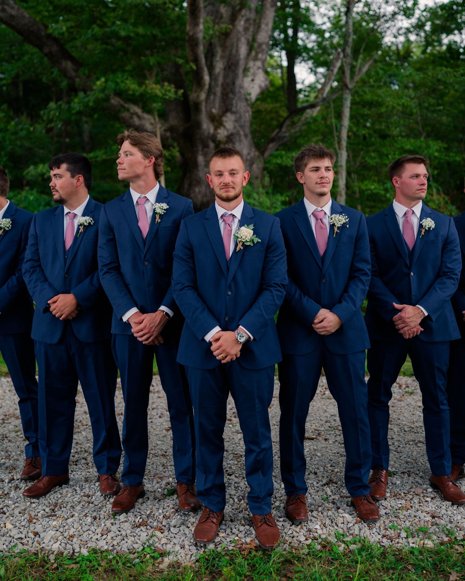Groomsmen in matching navy blue suits with pink ties standing in formation on gravel path.