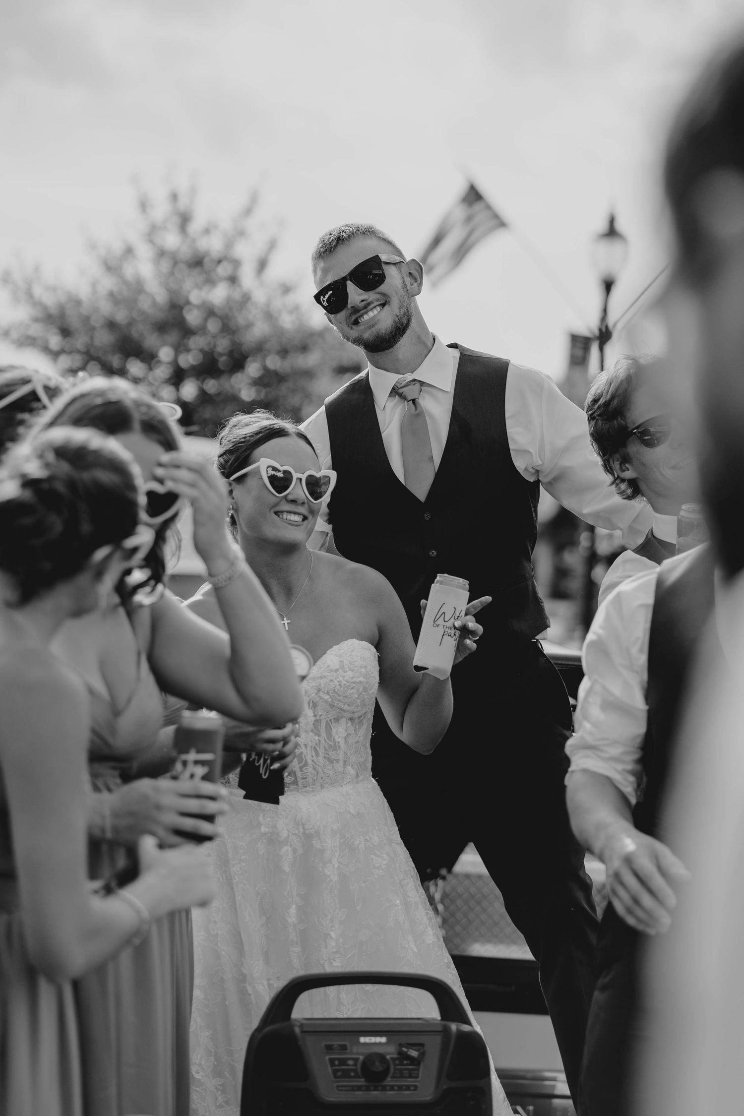 Black and white photo of bride and groom in sunglasses celebrating with wedding guests outdoors.