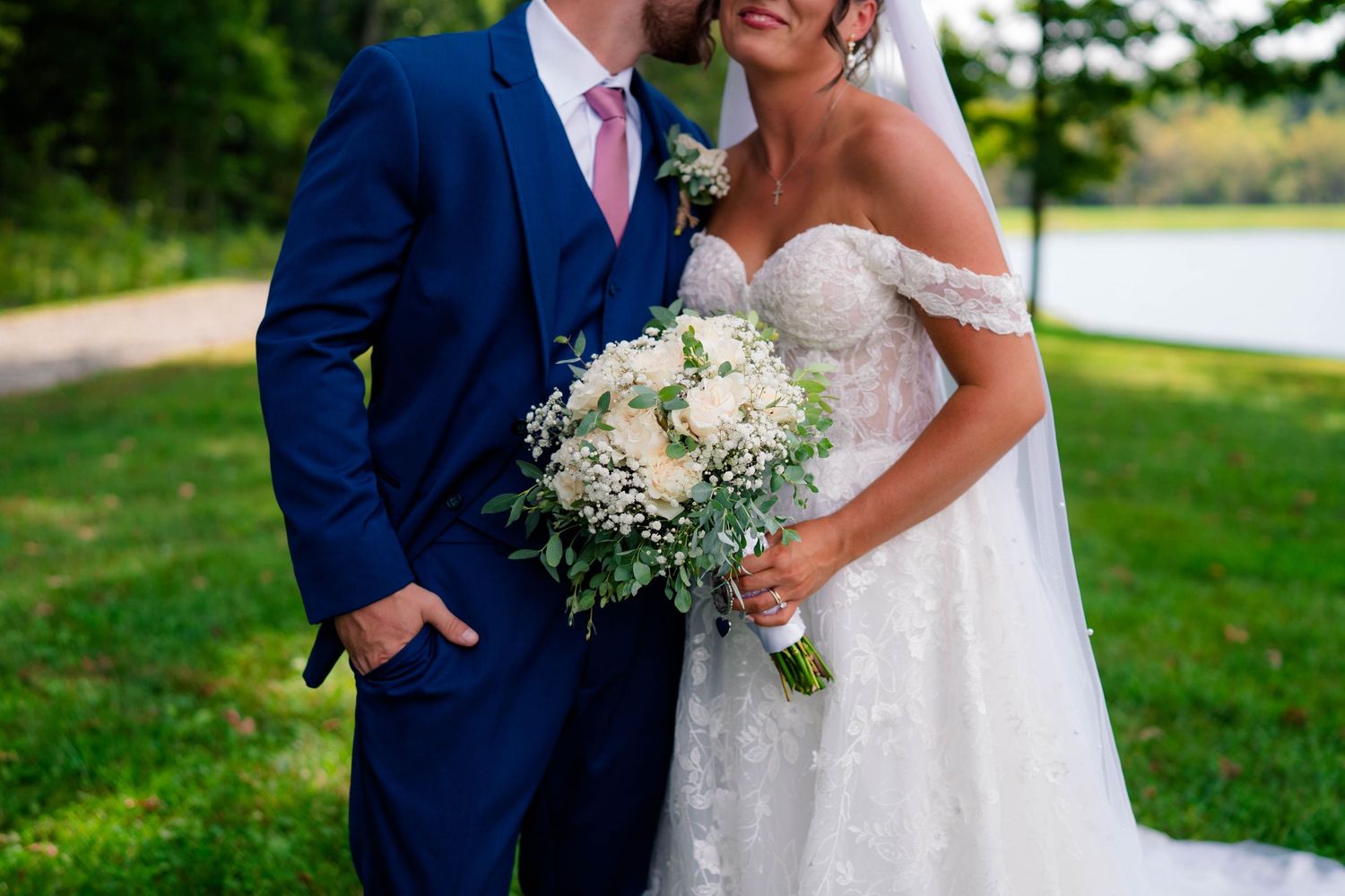 Bride and groom pose outdoors near a lake, she holds a white floral bouquet, he wears a navy suit with pink tie.