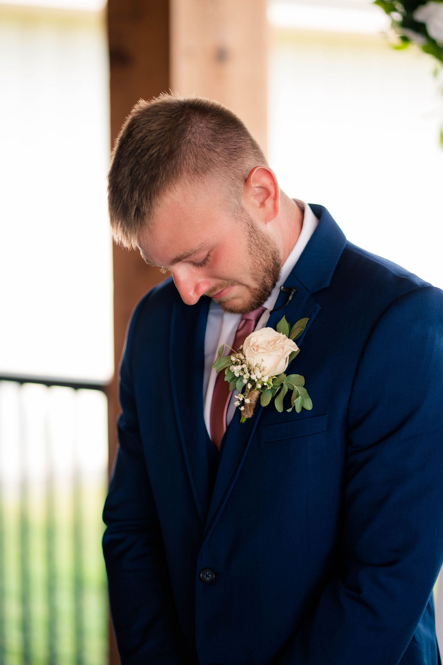Groom in navy suit with floral boutonniere bows head during wedding ceremony.