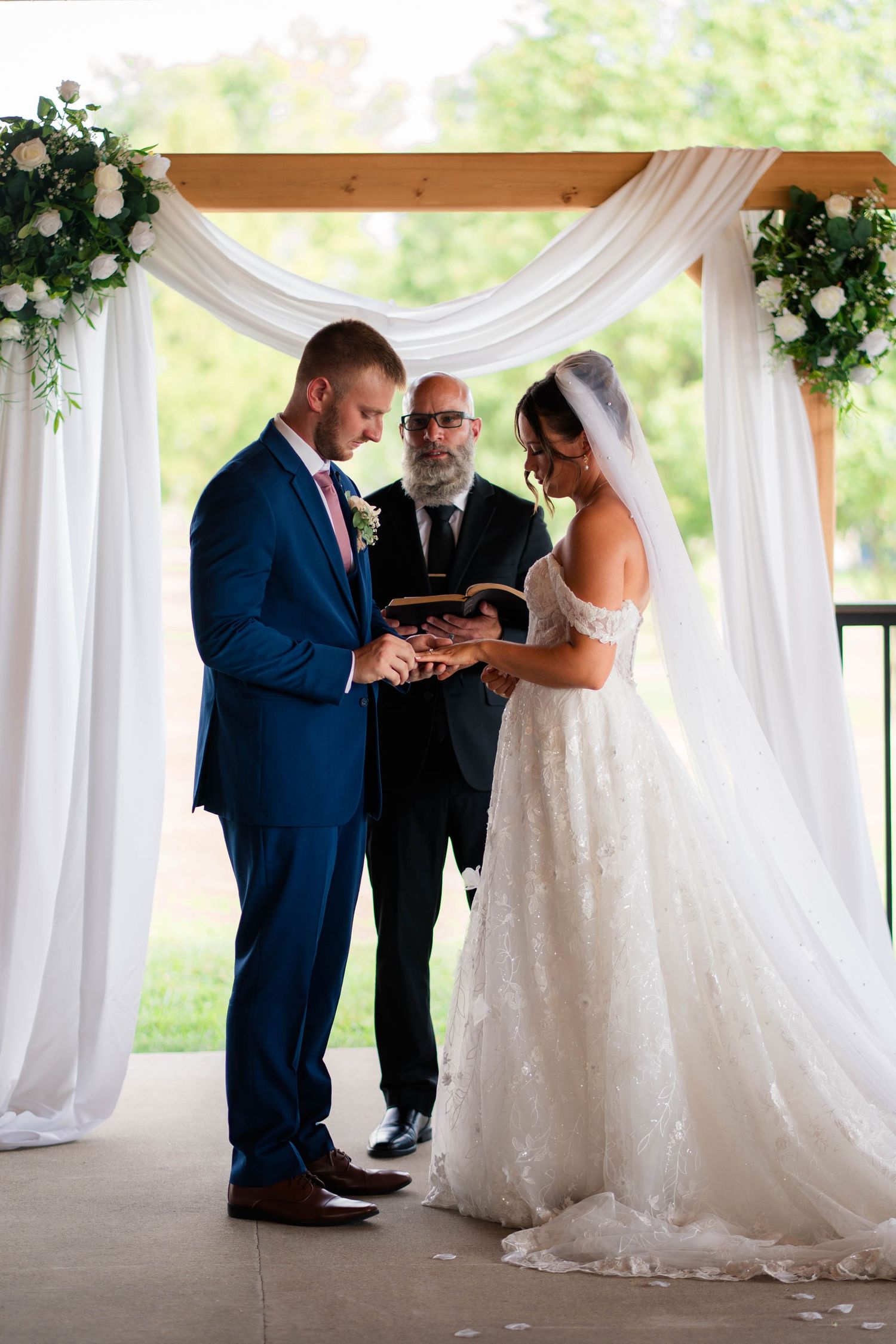 Bride and groom exchanging rings at outdoor wedding ceremony under white draped arch with floral arrangements.