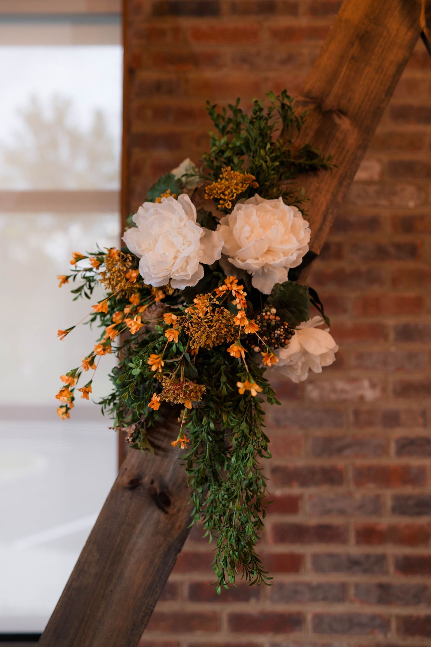Close-up of white peonies and orange wildflowers arranged on a rustic wooden triangular wedding arch.