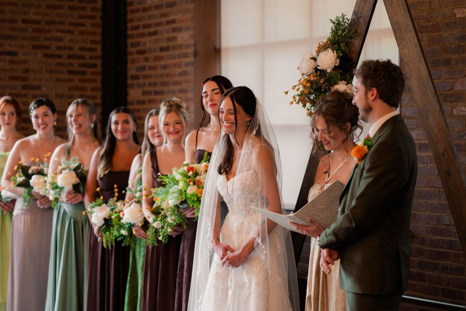 Bride laughing with groom and bridesmaids during wedding ceremony at rustic brick venue.