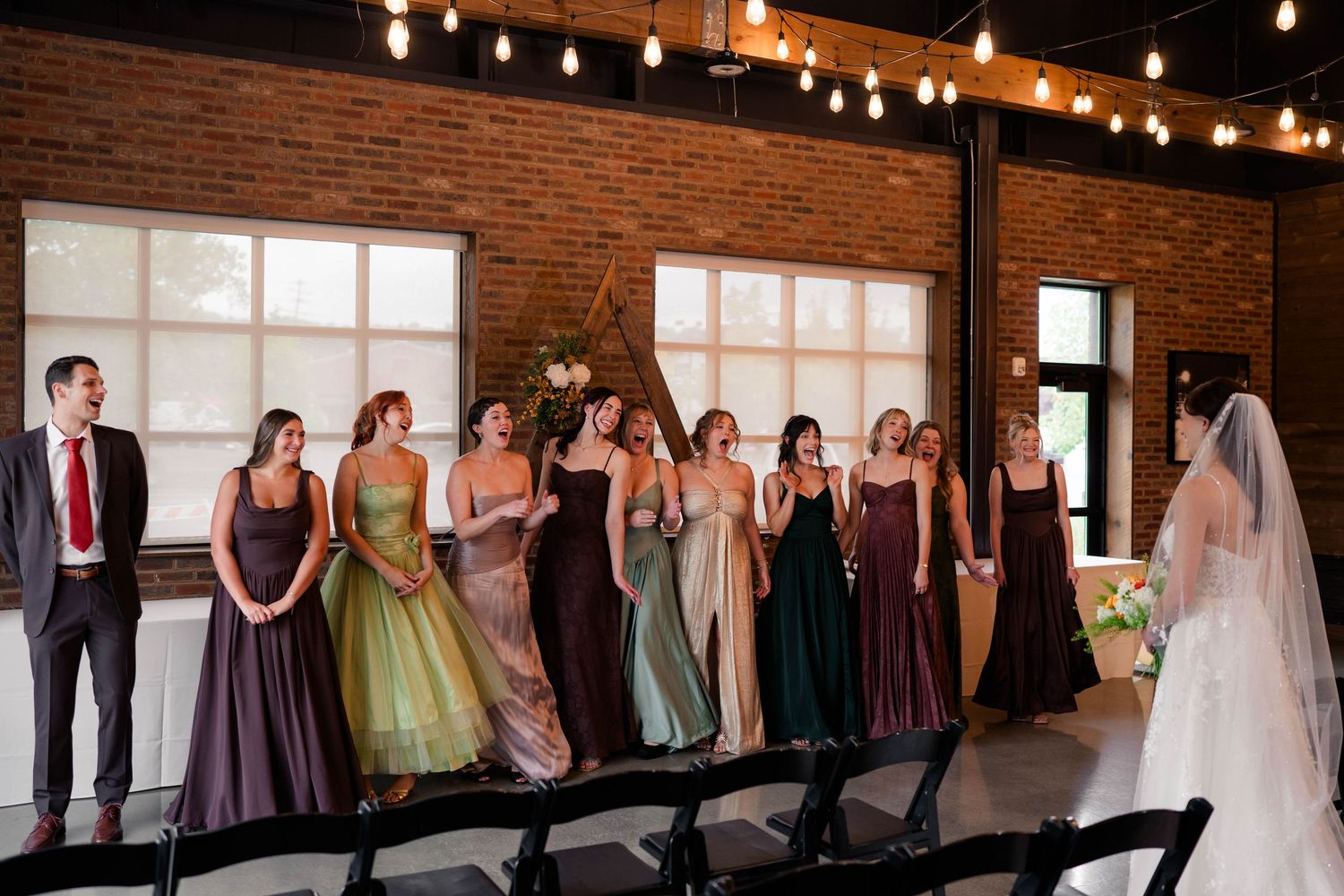 Bridesmaids in mismatched jewel-tone gowns stand smiling at a bride during an indoor wedding ceremony with Edison bulb lighting.
