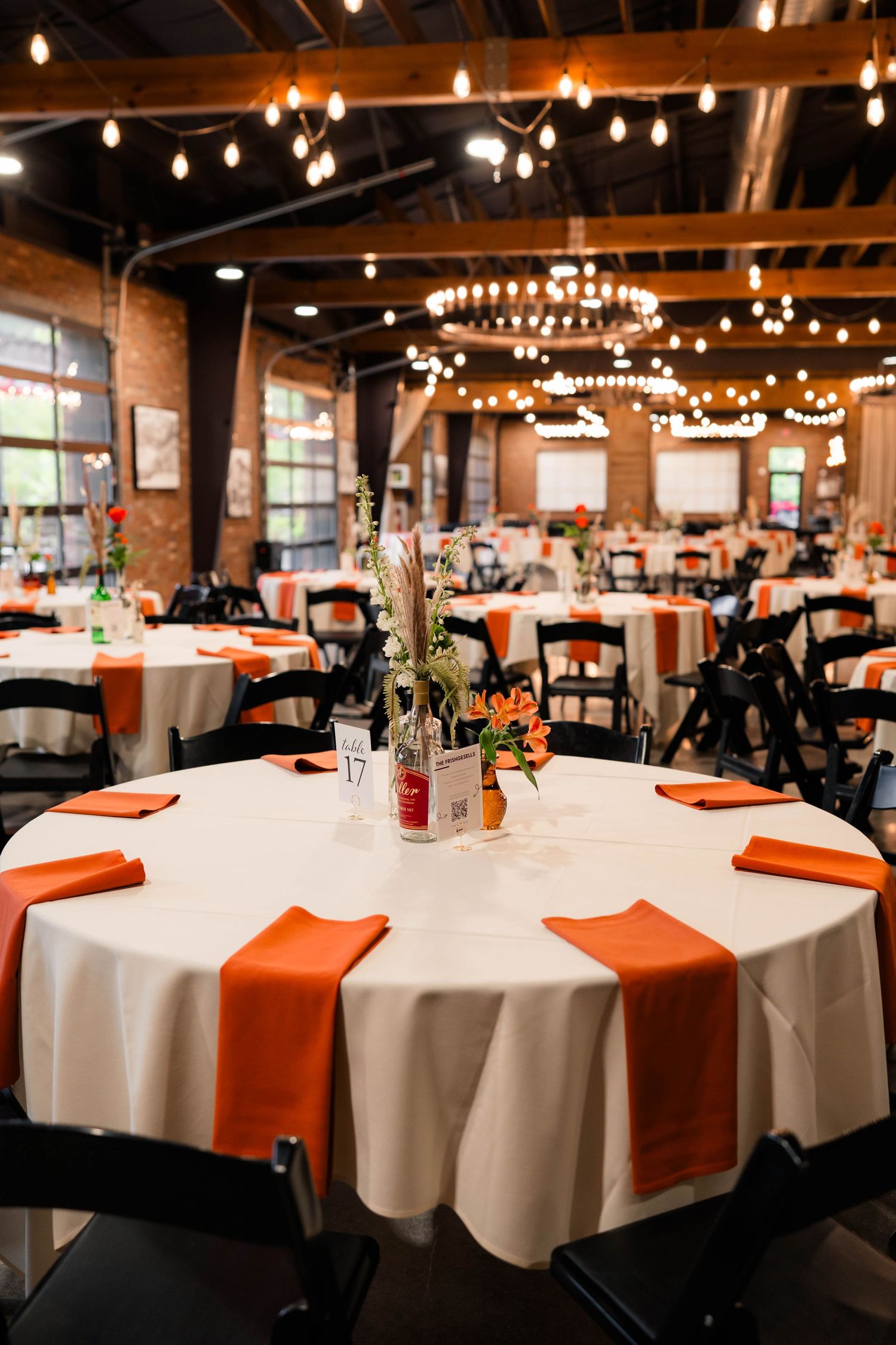 Elegant wedding reception hall with round tables, orange napkins, string lights, and floral centerpieces.