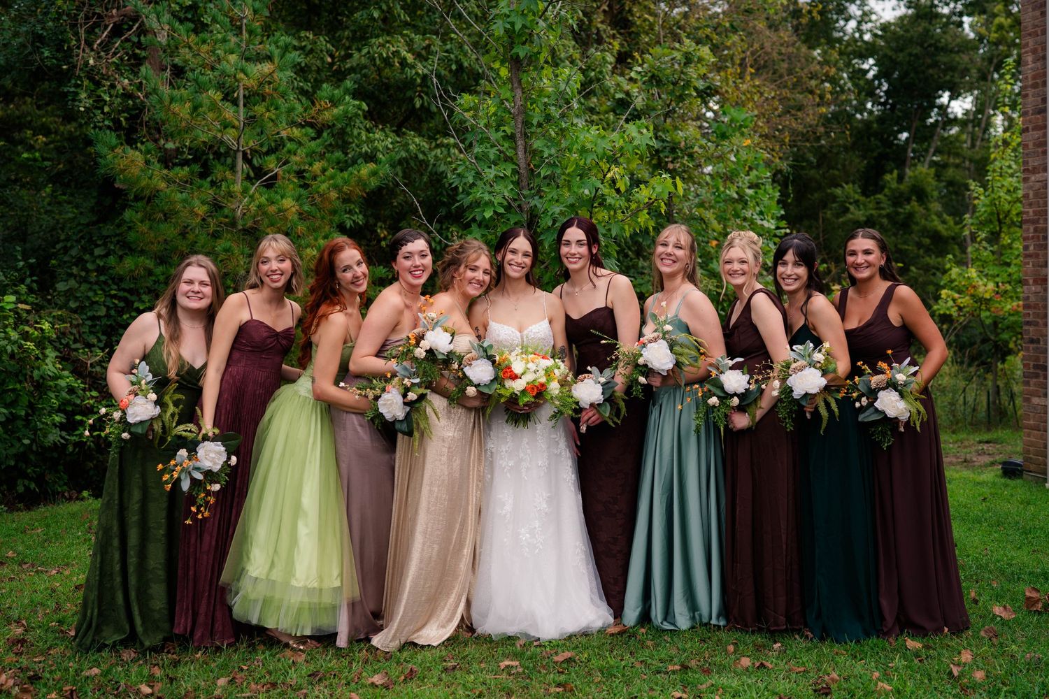 Bride poses with eight bridesmaids in mismatched green and brown gowns holding floral bouquets outdoors.