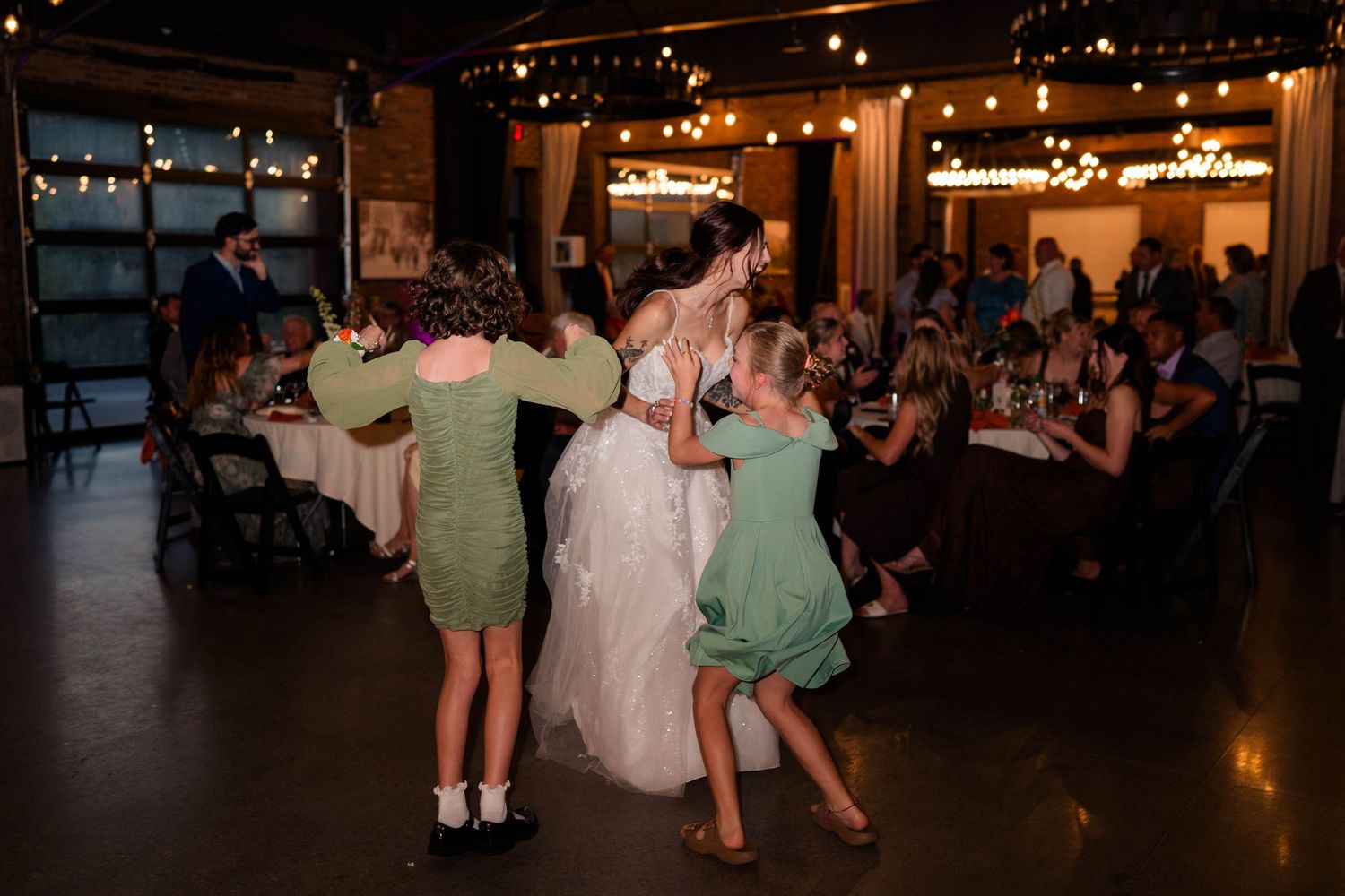 Bride dances joyfully with young flower girls in green dresses during wedding reception celebration.