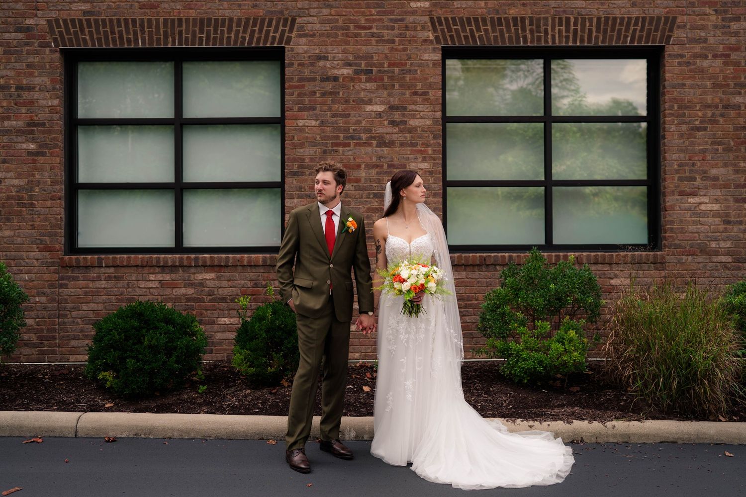Wedding couple stands before brick building, bride in lace gown holding colorful bouquet, groom in olive suit.
