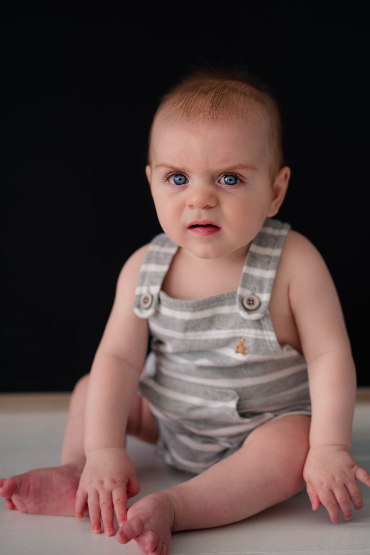 Blue-eyed baby in striped gray overalls sitting on white surface against black background.