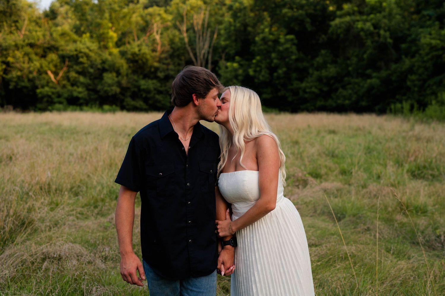 Couple sharing a romantic kiss in a golden meadow with lush green trees in the background.