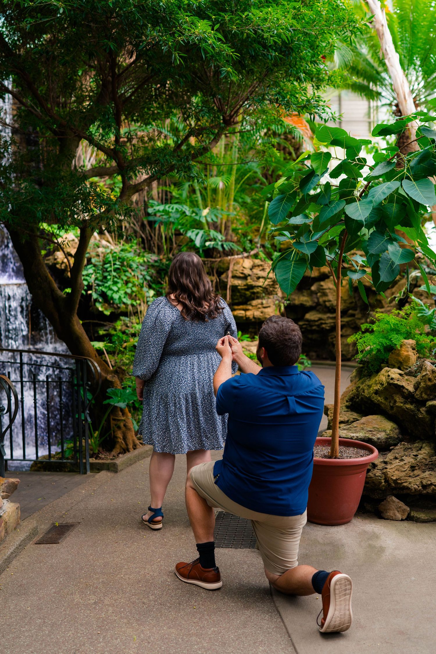 Man on one knee proposing to woman in floral dress near waterfall and tropical plants indoors.