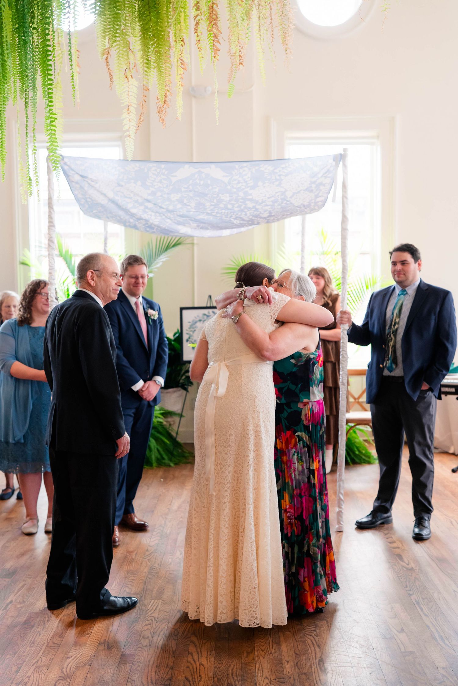 Bride in lace gown embraces guest under chuppah during Jewish wedding ceremony with attendants.