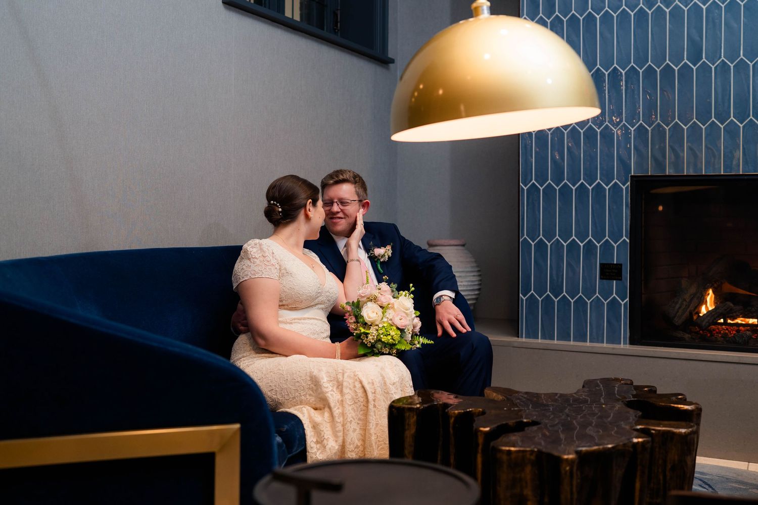 Bride and groom sitting together on a navy sofa under a gold pendant light near a tiled fireplace.