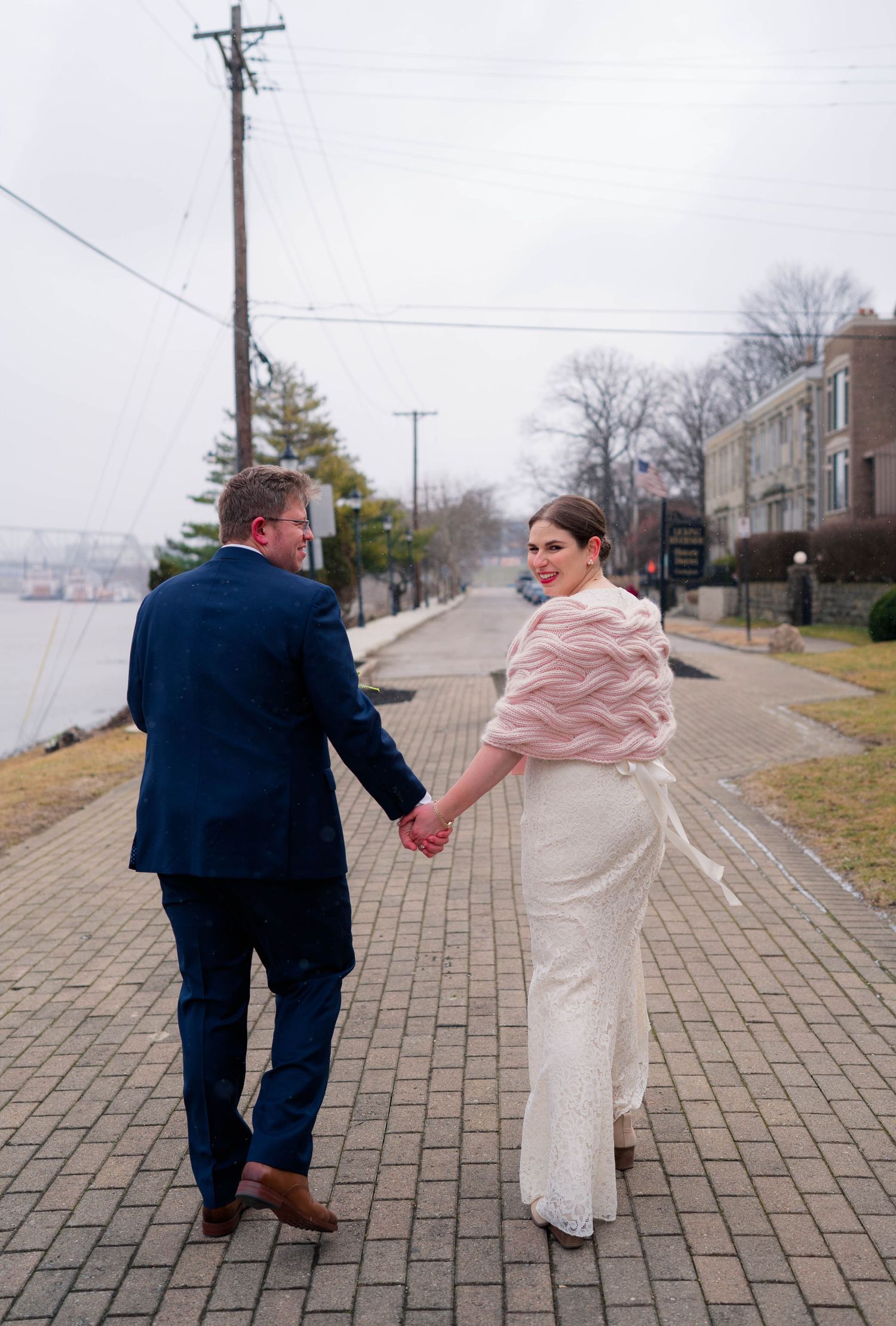 Bride and groom walking hand in hand along a brick riverside path, bride looking back at the camera.