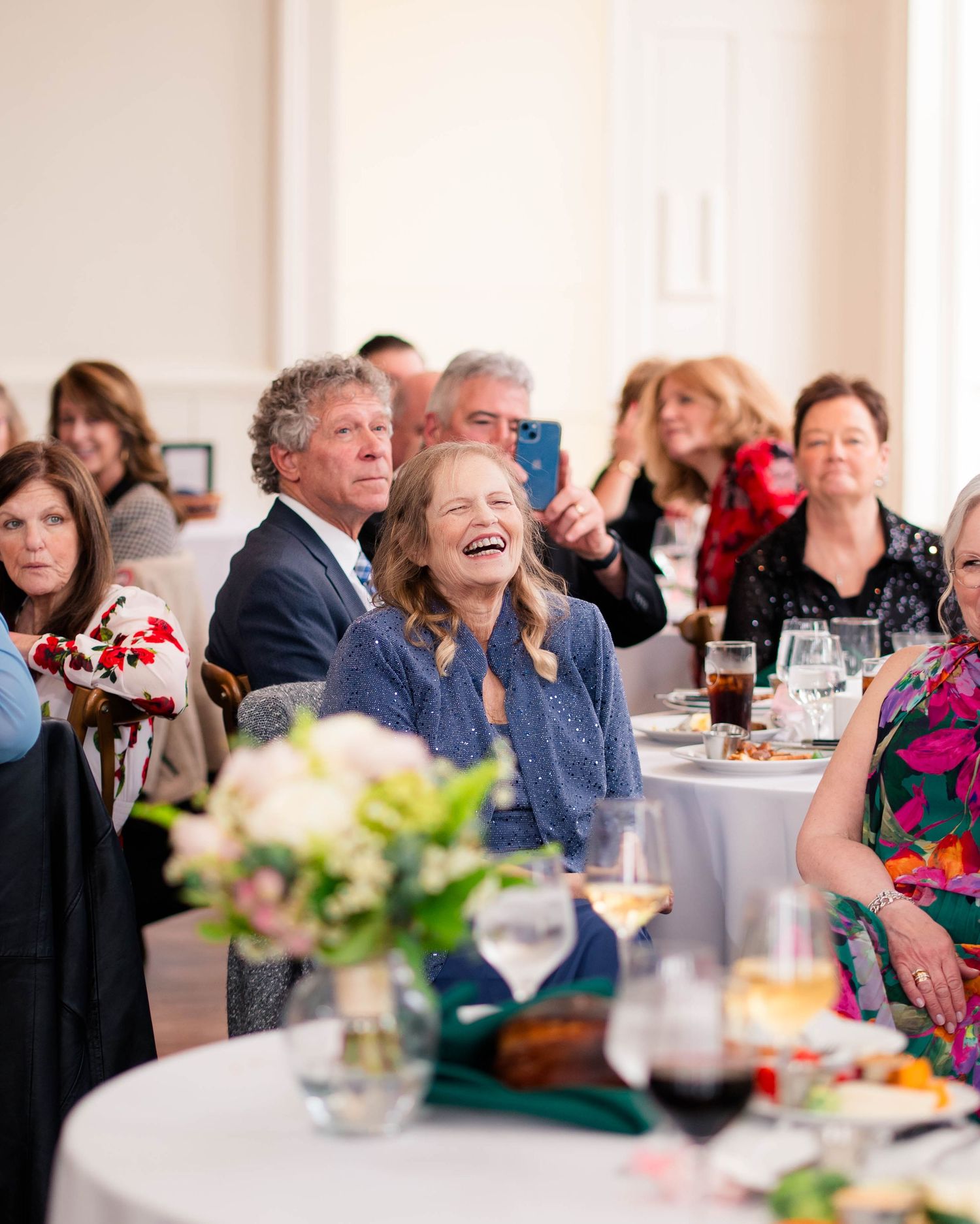 Wedding guests laugh and record speeches on phones during elegant indoor reception dinner.