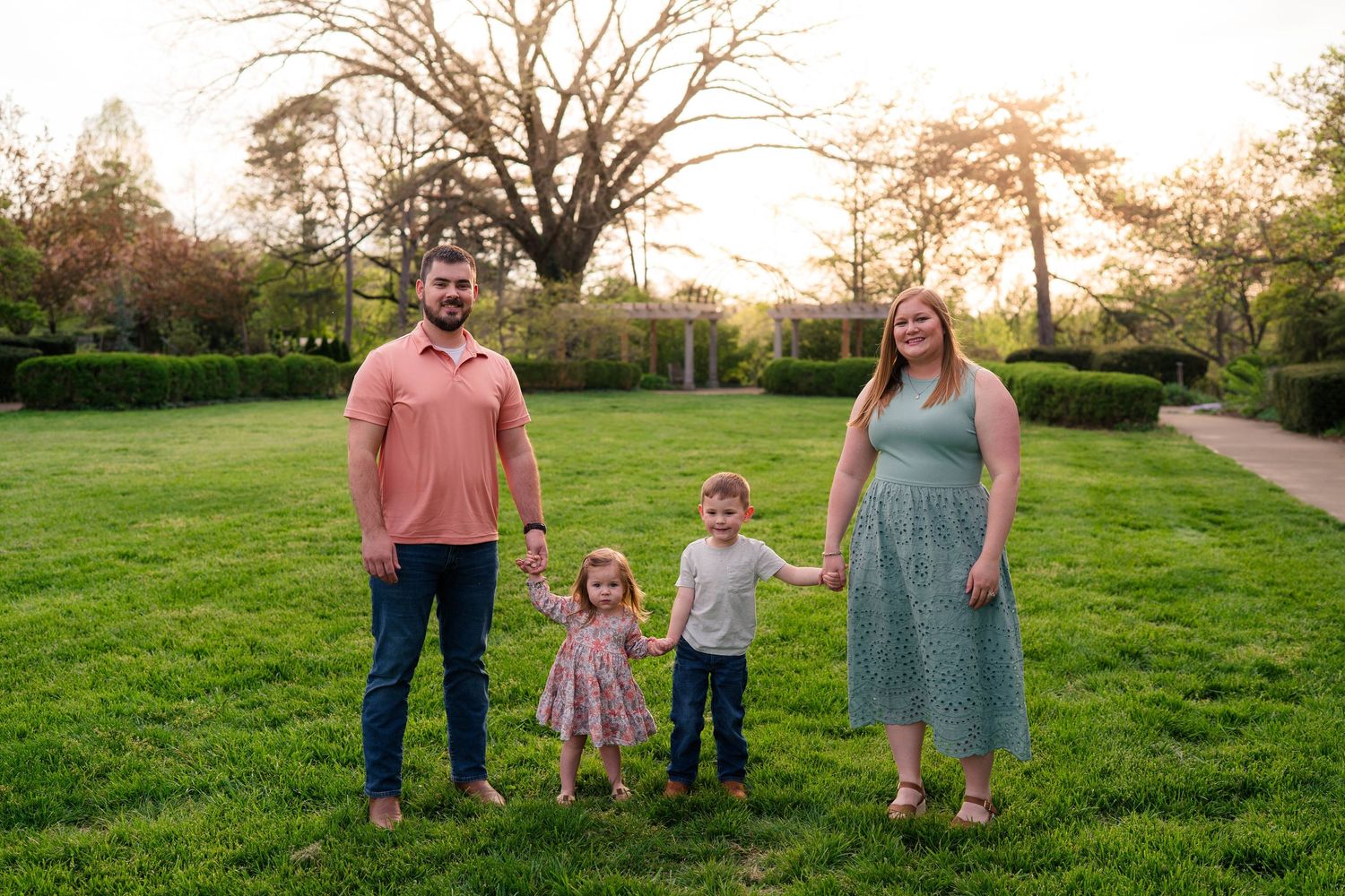 Family of four holding hands on green grass at sunset in a scenic park setting.