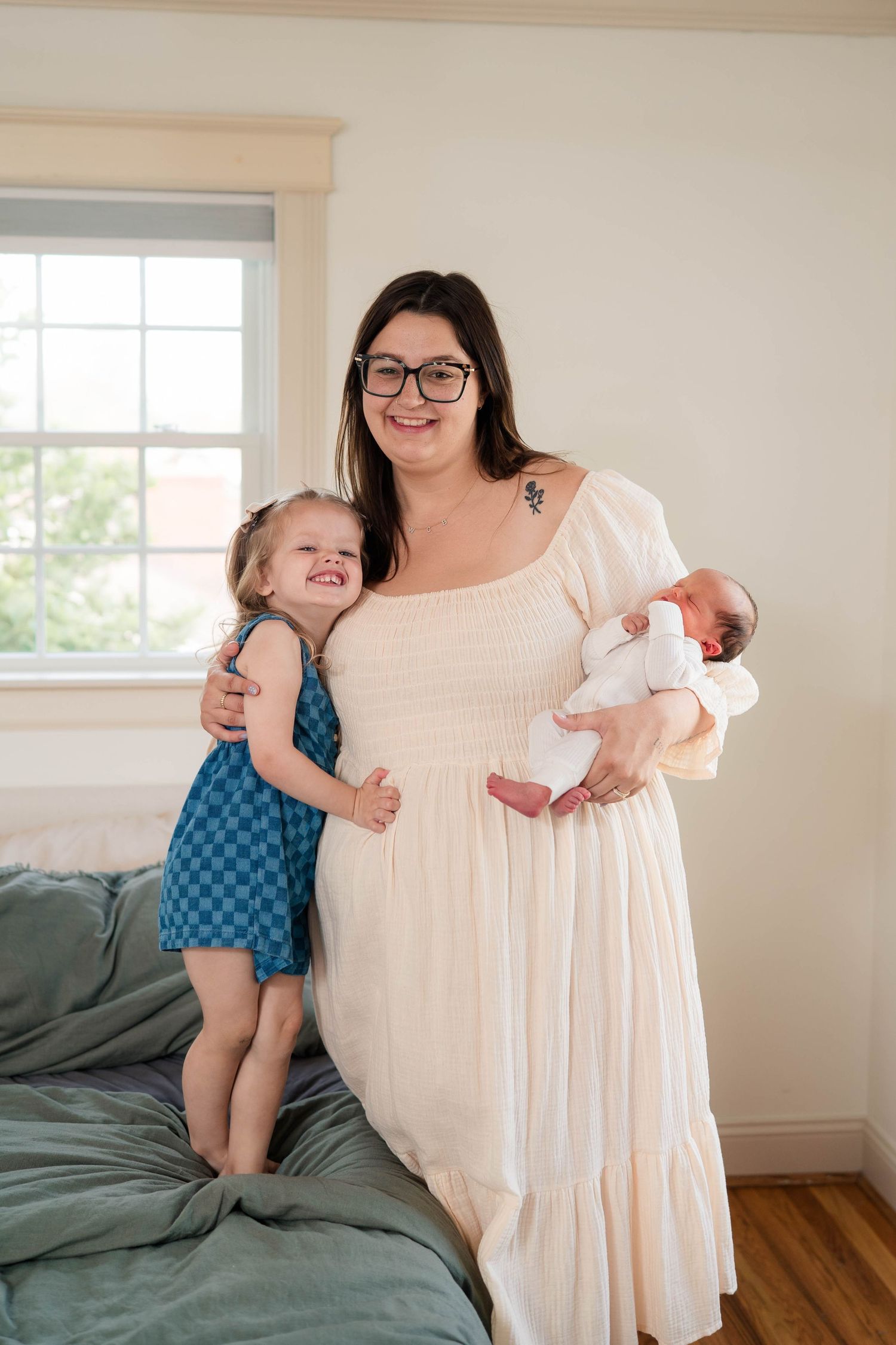 Mother in white dress holds newborn baby while toddler daughter in blue dress hugs her side.