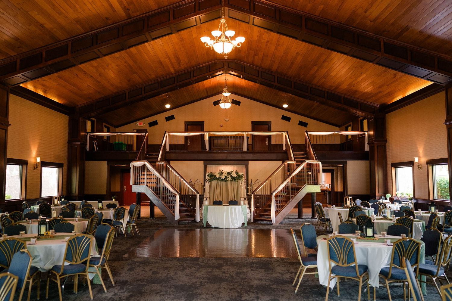 Elegant wedding reception hall with wooden ceiling, round tables, and dual staircases.
