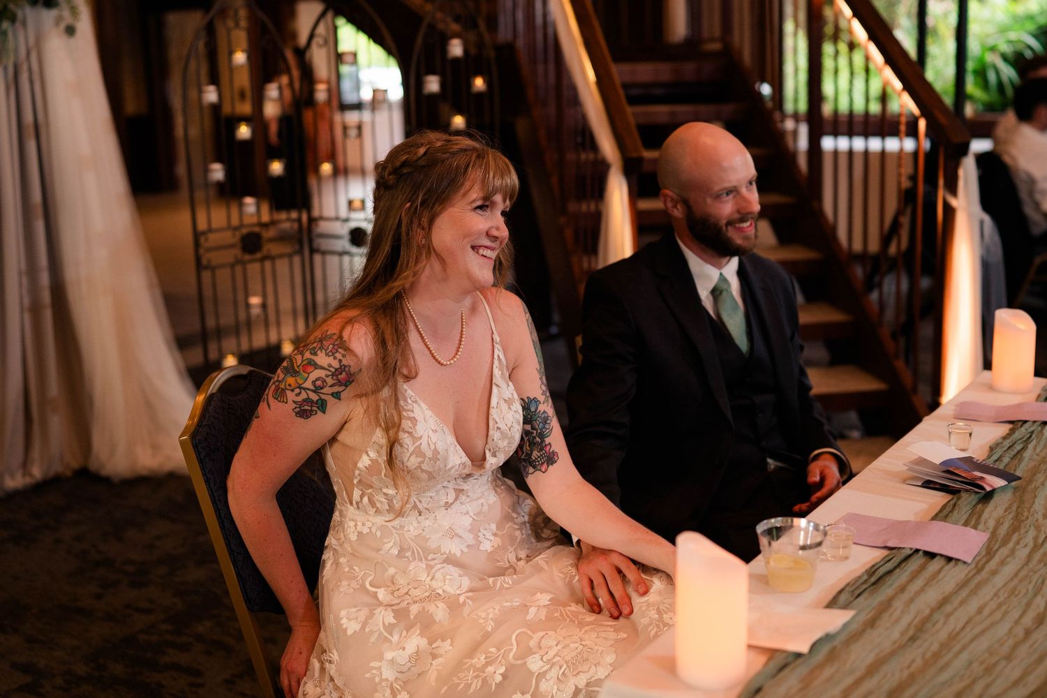 Laughing bride in lace gown and groom sit together at sweetheart table during wedding reception.