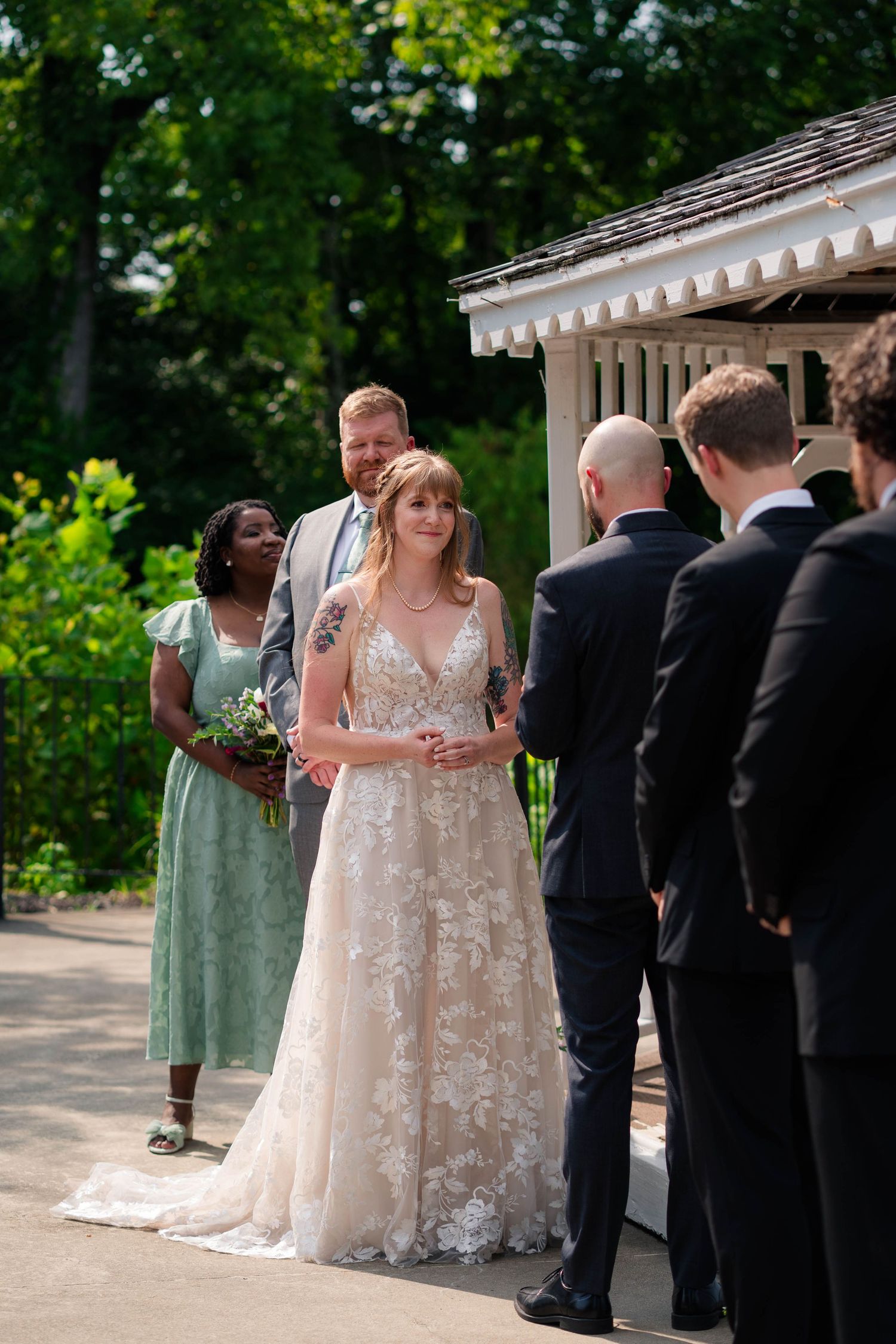A bride in a floral lace wedding gown stands at the altar during an outdoor garden ceremony.
