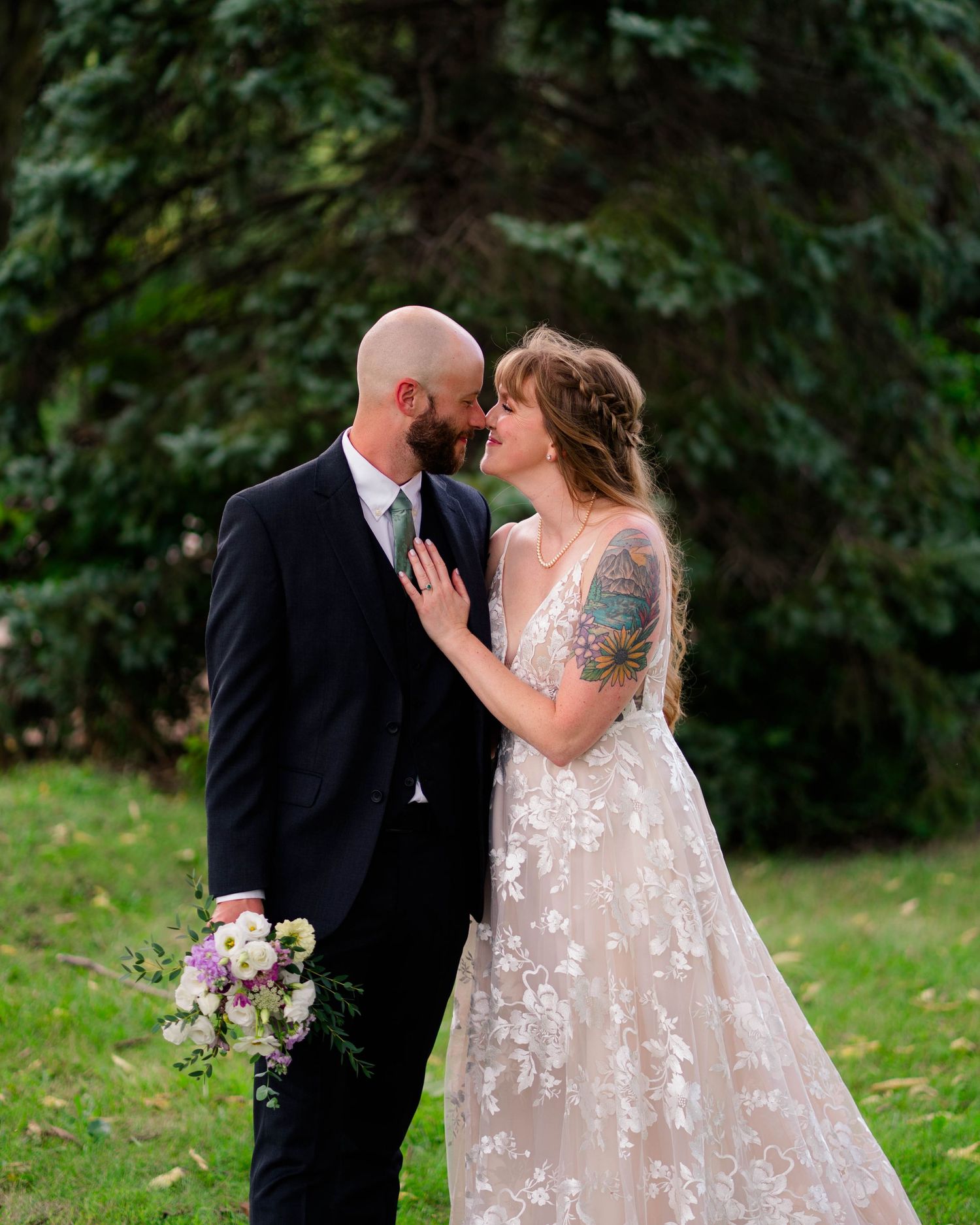 Tattooed bride and bald groom gaze at each other in lush green garden setting.