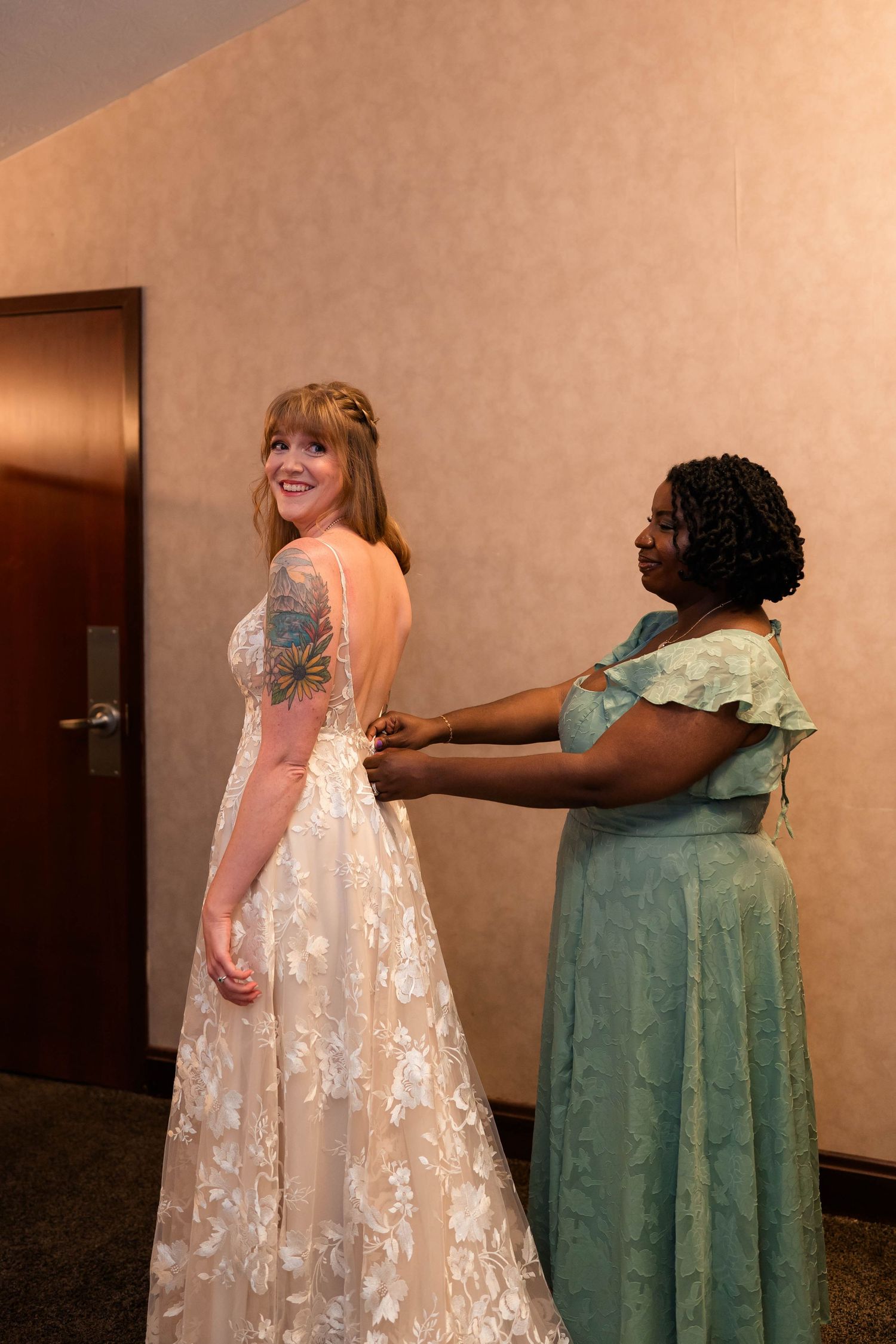 A bridesmaid in teal helps a bride in a floral lace backless gown get dressed before her wedding ceremony.