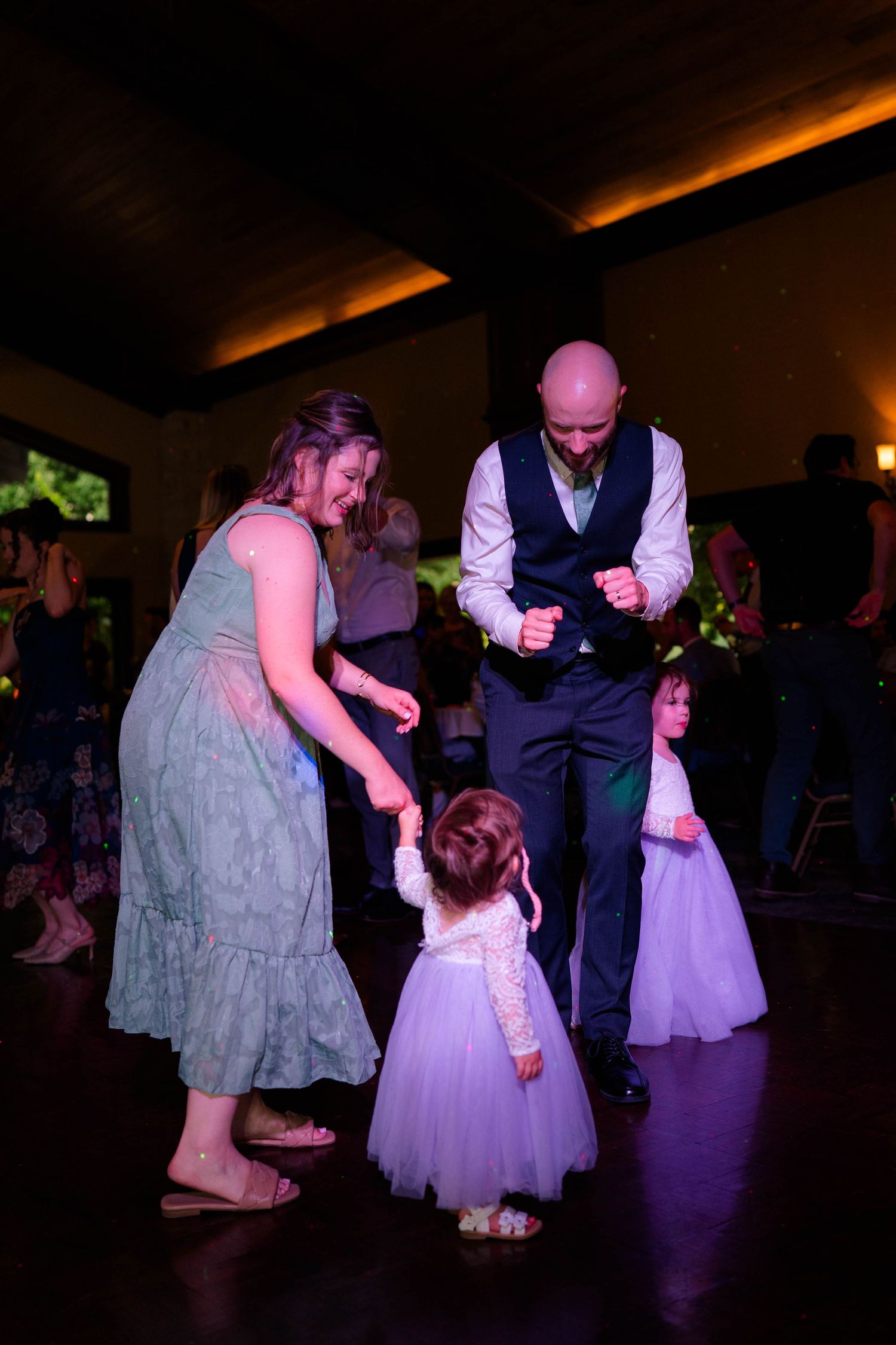 Bald groom dances joyfully with a woman in green and two little flower girls in lavender dresses at the reception.