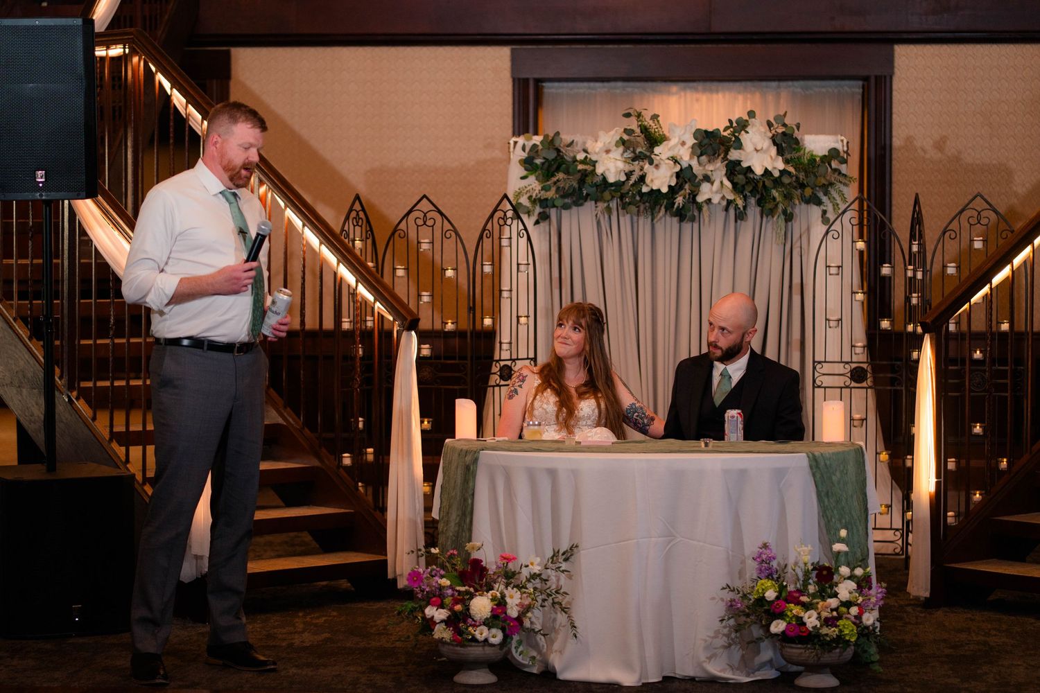 Groomsman delivers heartfelt speech at microphone while bride and groom sit at floral sweetheart table.