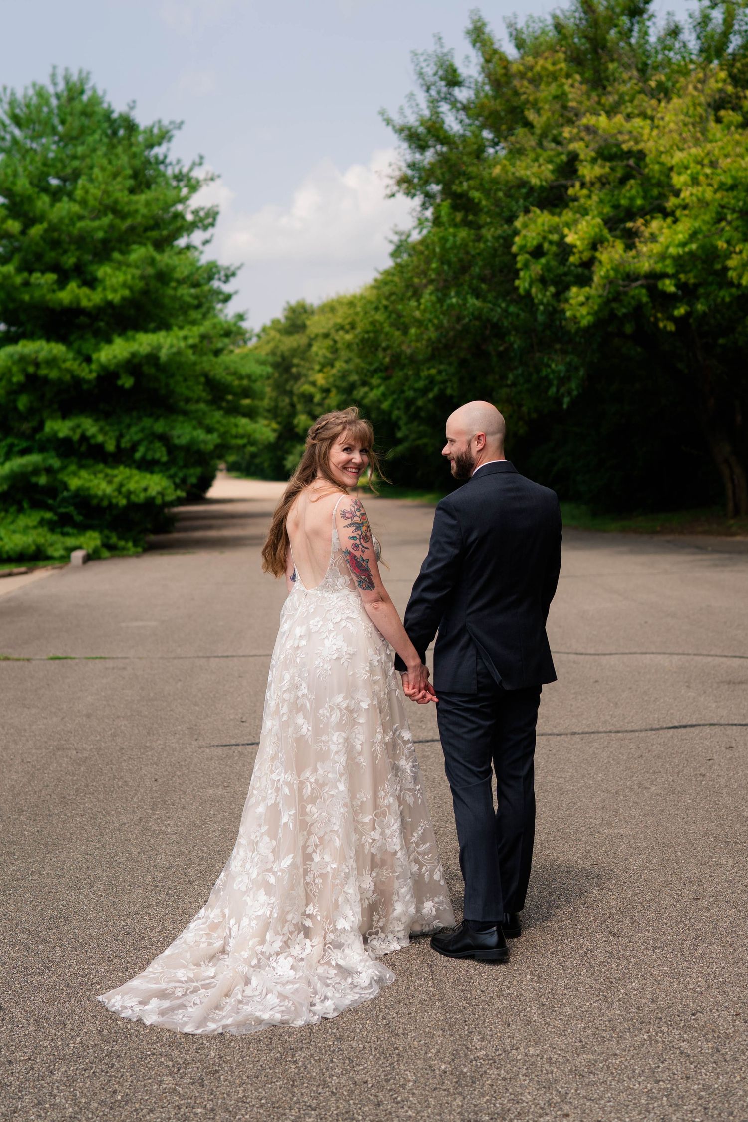 Bride in floral lace gown with train looks back smiling while holding hands with bald groom outdoors.