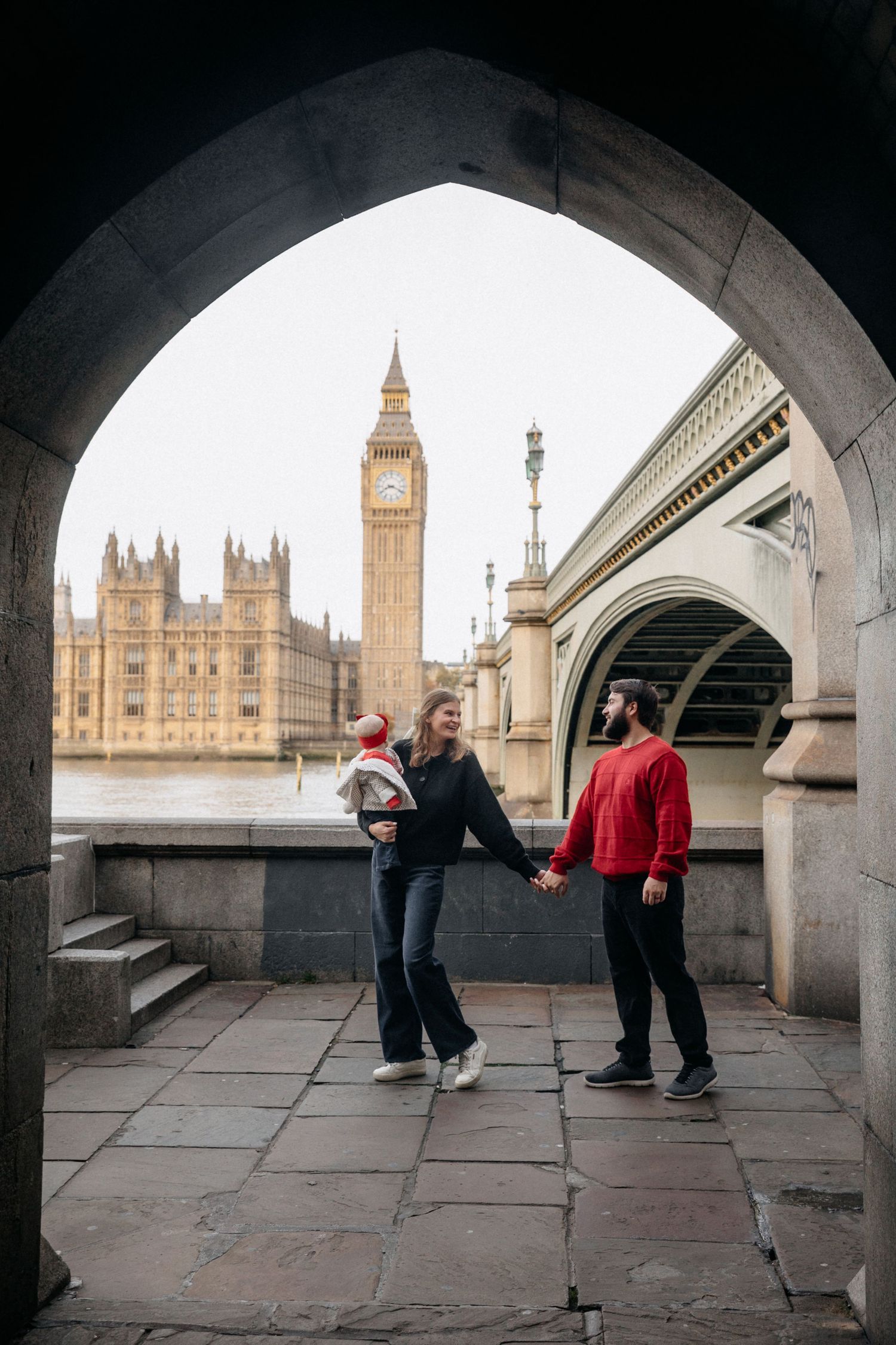Young family with baby standing under an arch with Big Ben and Westminster Bridge in London.