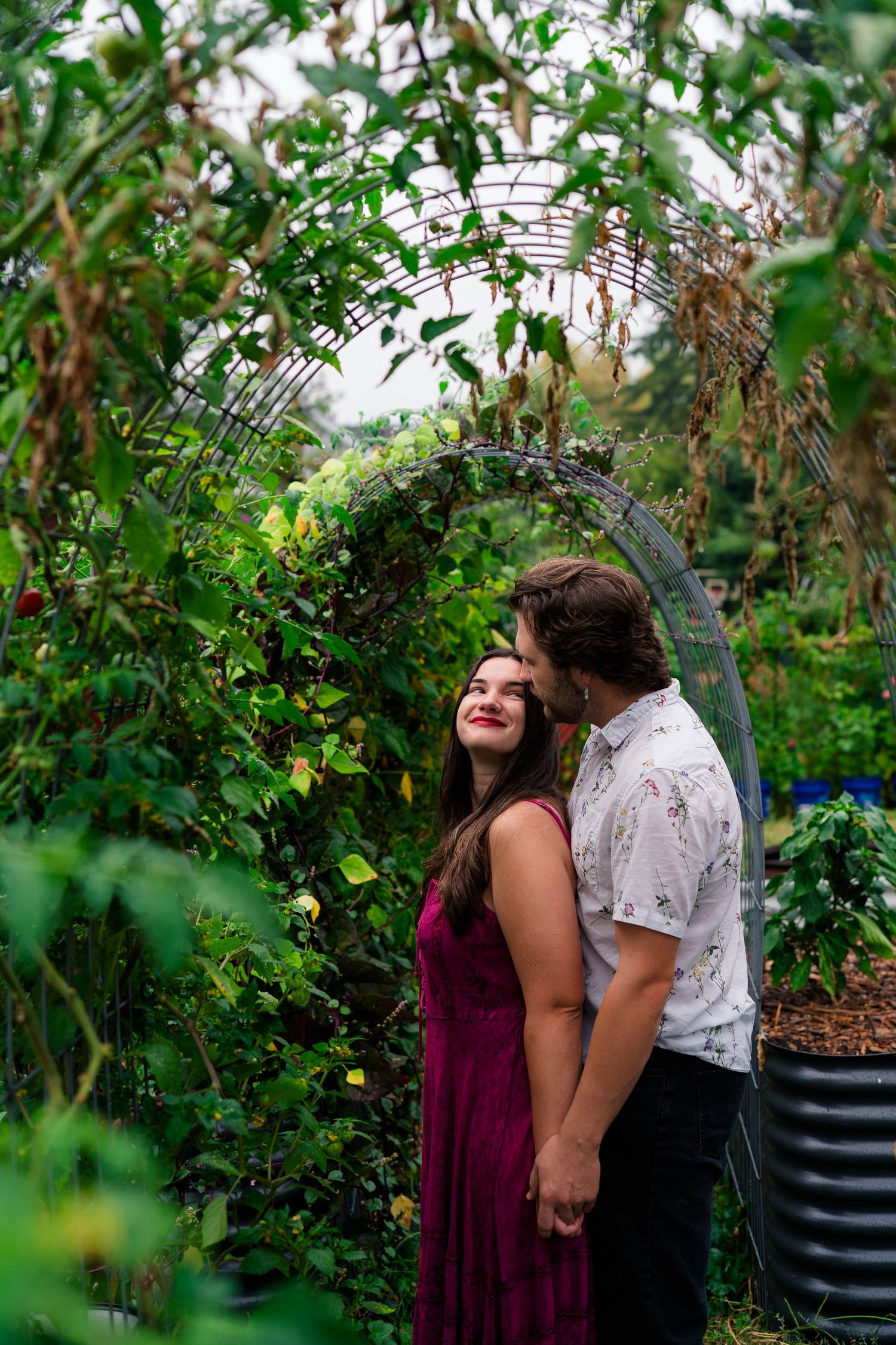 Couple sharing a tender kiss under a lush green garden arch during outdoor engagement session.