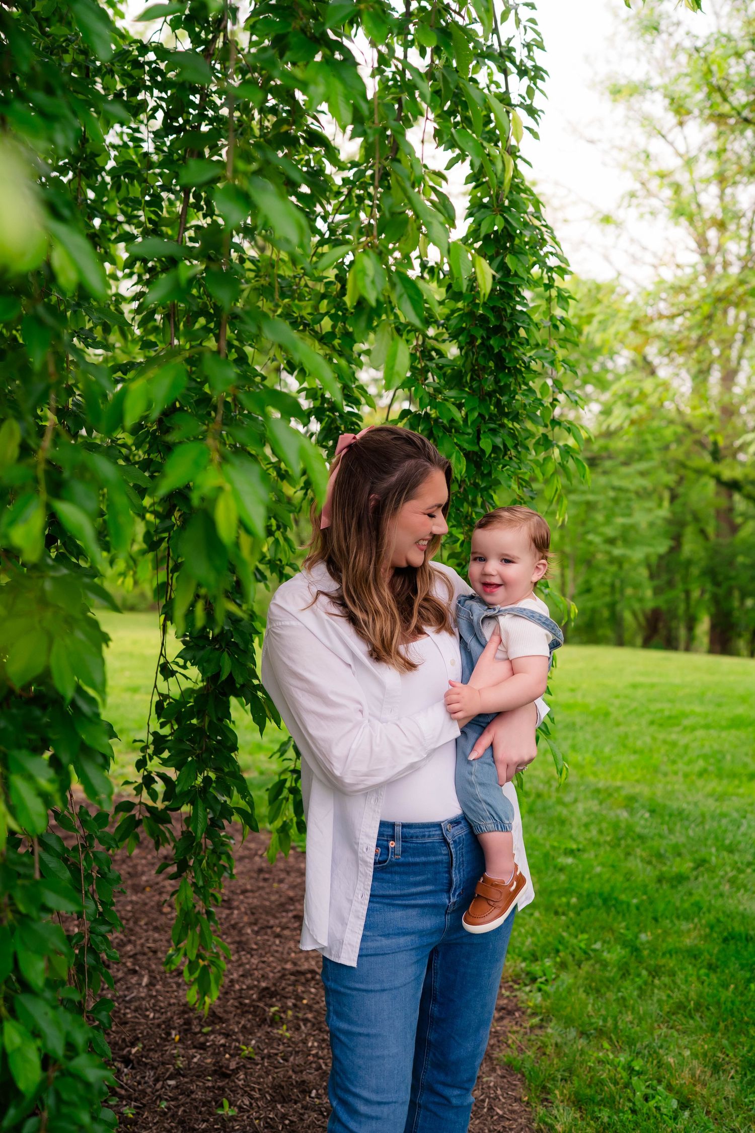 A mother in a white shirt and jeans holds her smiling baby outdoors under lush green tree branches.