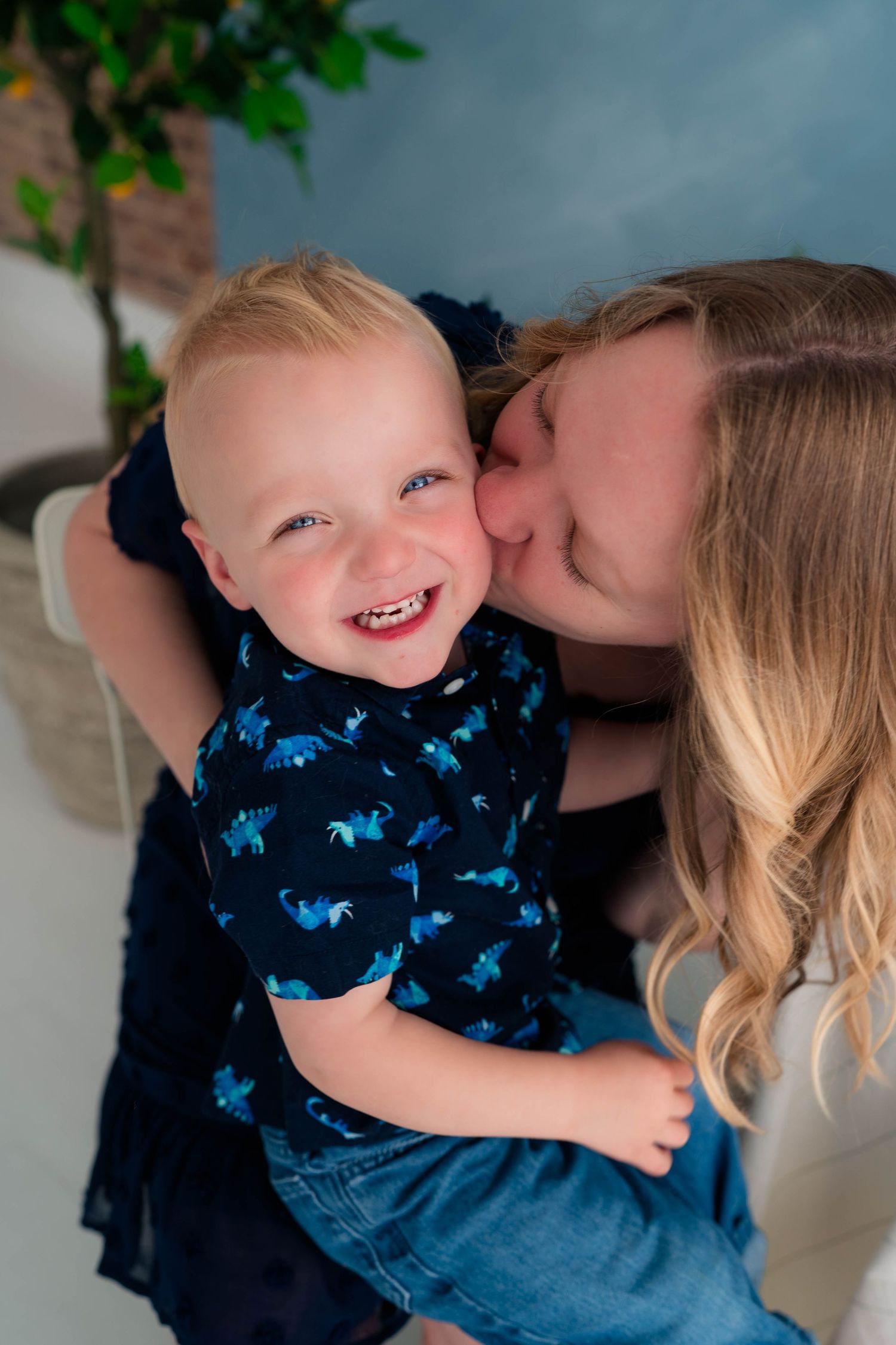 A joyful toddler in a dinosaur shirt laughs as a woman with long hair gives him a loving kiss on the cheek.